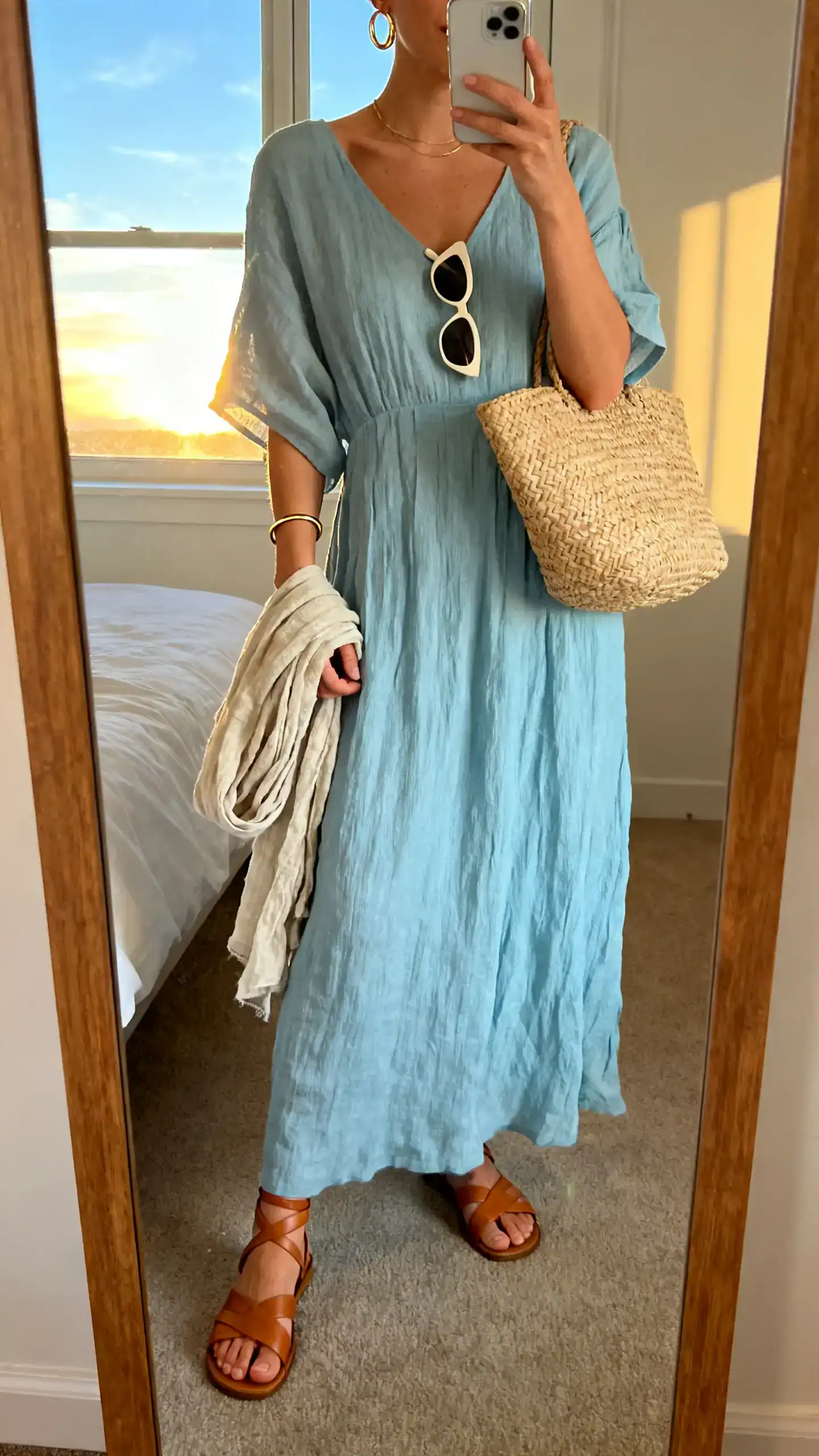 iPhone mirror selfie of a woman wearing a flowy linen midi dress in soft sky blue, tan leather strappy sandals, a raffia tote, delicate gold hoops with a thin bangle, minimal cat-eye sunglasses, and a lightweight linen scarf draped over her arm, face not visible, cropped at neck, bedroom mirror, golden hour window light, iPhone photo quality.