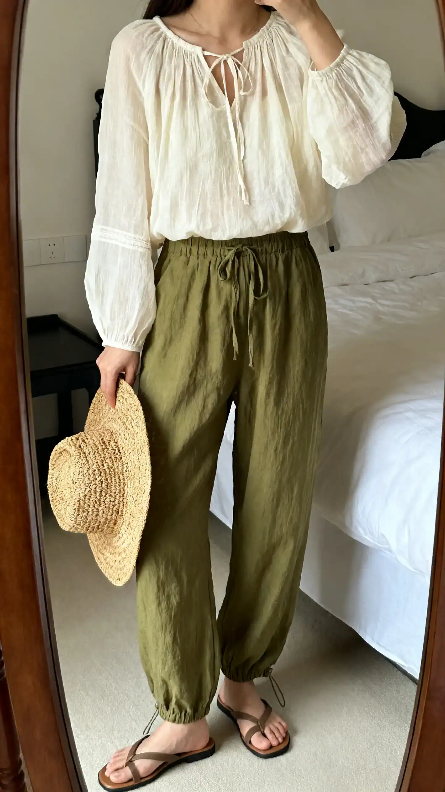iPhone mirror selfie of a woman wearing a gauzy cream peasant top with subtle ties at the neckline, relaxed linen drawstring pants in olive, slide sandals, woven straw hat held at side, face not visible, cropped at shoulders, bedroom mirror, soft natural light, iPhone photo quality