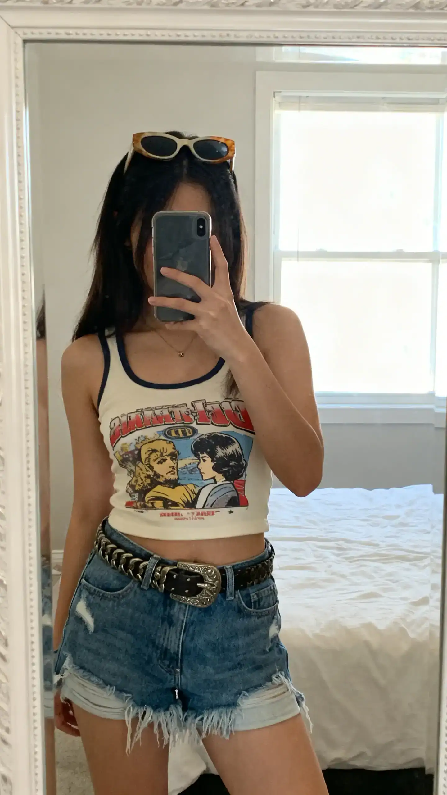 iPhone mirror selfie of a woman wearing a graphic baby tank with frayed denim cutoffs, chunky belt, and retro sunglasses pushed up on her head, face not visible, cropped at shoulders, bedroom mirror, bright window light, iPhone photo quality.