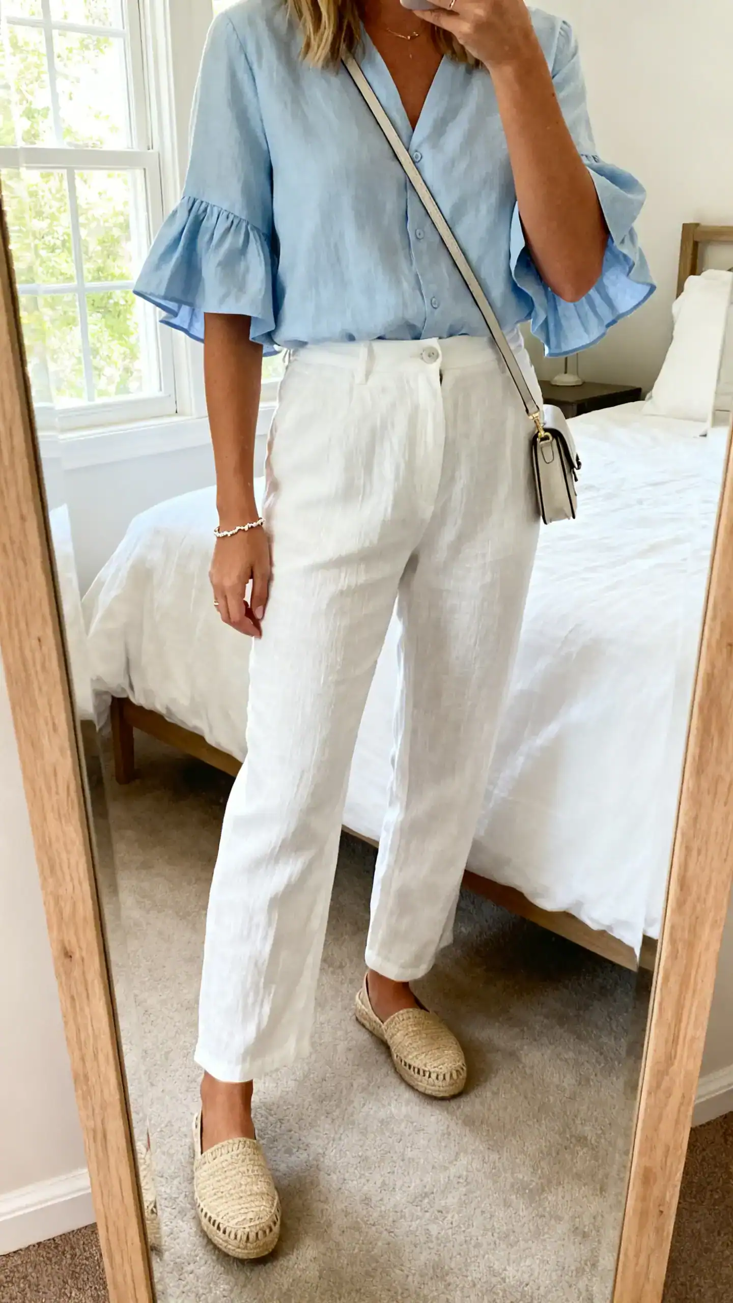 iPhone mirror selfie of a woman wearing a light blue flutter-sleeve blouse tucked into white linen trousers, espadrille flats, delicate bracelet, small crossbody bag, face not visible, cropped at shoulders, bedroom mirror, natural window light, iPhone photo quality