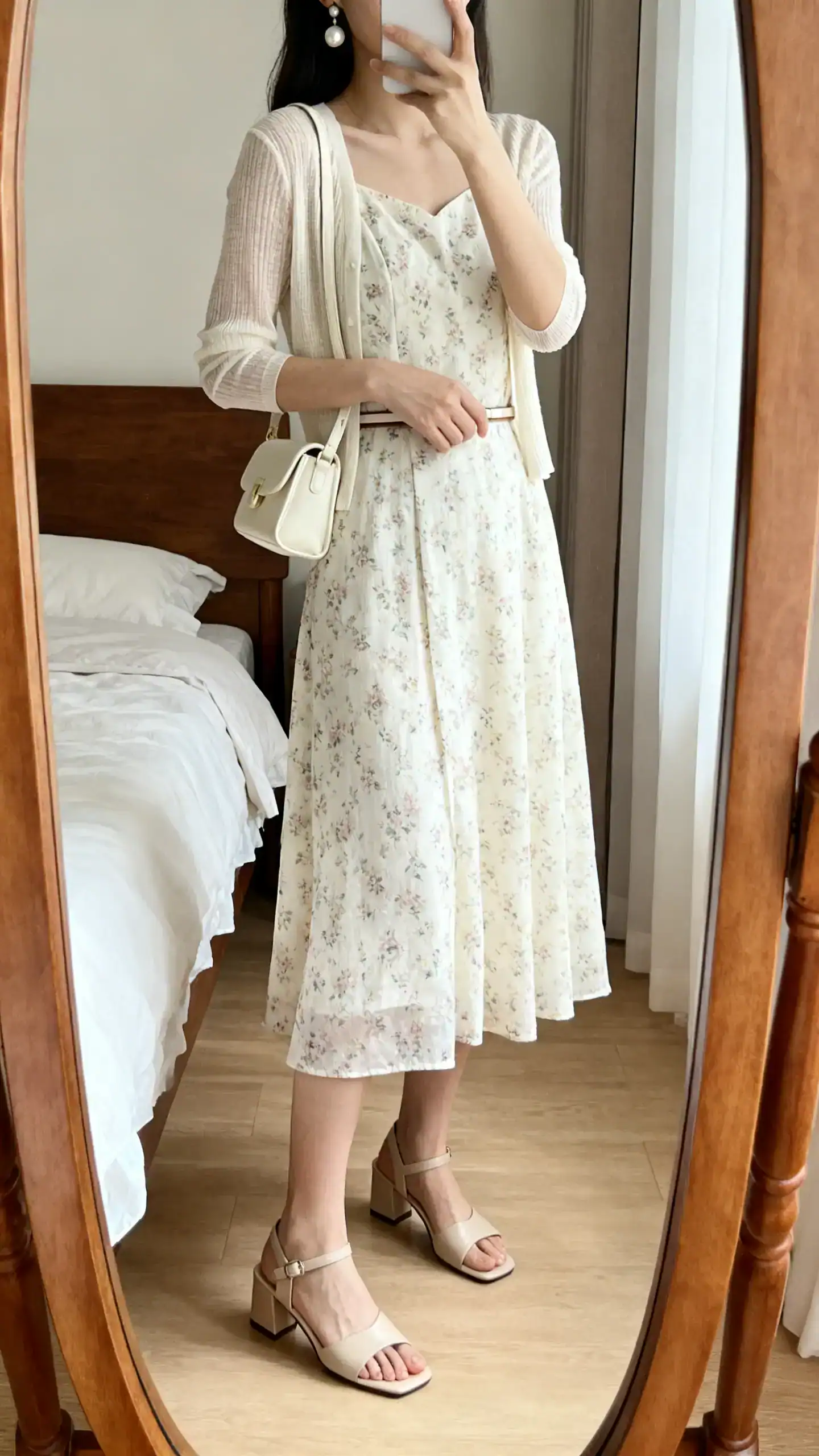 iPhone mirror selfie of a woman wearing a light floral tea dress just-below-the-knee, block-heel neutral leather sandals, a small cream shoulder bag, pearl drop earrings, a fine cardigan over shoulders, and a thin waist belt, face not visible, cropped at neck, bedroom mirror, natural daylight, iPhone photo quality.
