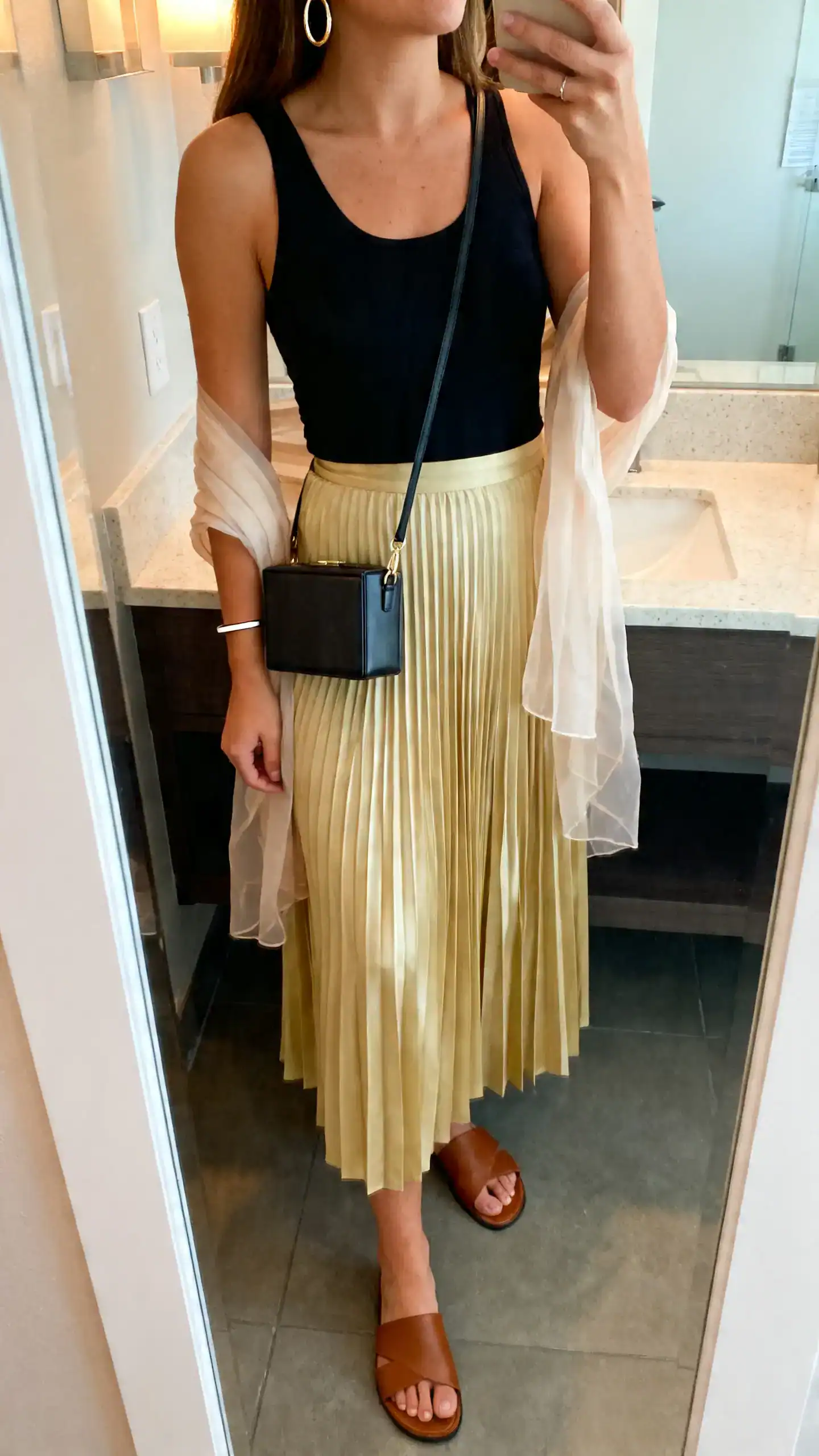iPhone mirror selfie of a woman wearing a light pleated midi skirt in soft gold, a fitted black tank, minimal leather slides, a boxy mini crossbody, medium hoop earrings, slim cuff bracelet, and a sheer wrap over her shoulders; face not visible, cropped at shoulders, bathroom mirror, indoor lighting, iPhone photo quality.