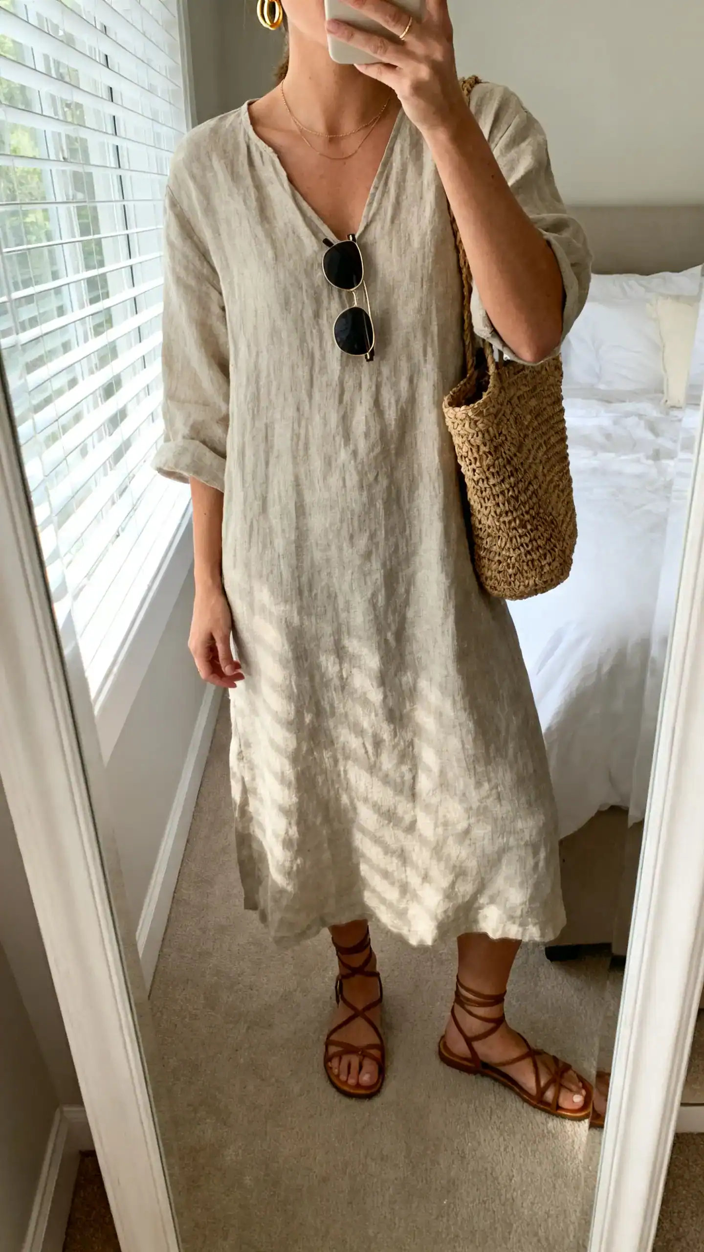 iPhone mirror selfie of a woman wearing a linen midi dress with strappy sandals, woven tote, delicate gold hoops, and slim sunglasses, face not visible, cropped at neck, bedroom mirror, morning window light, iPhone photo quality