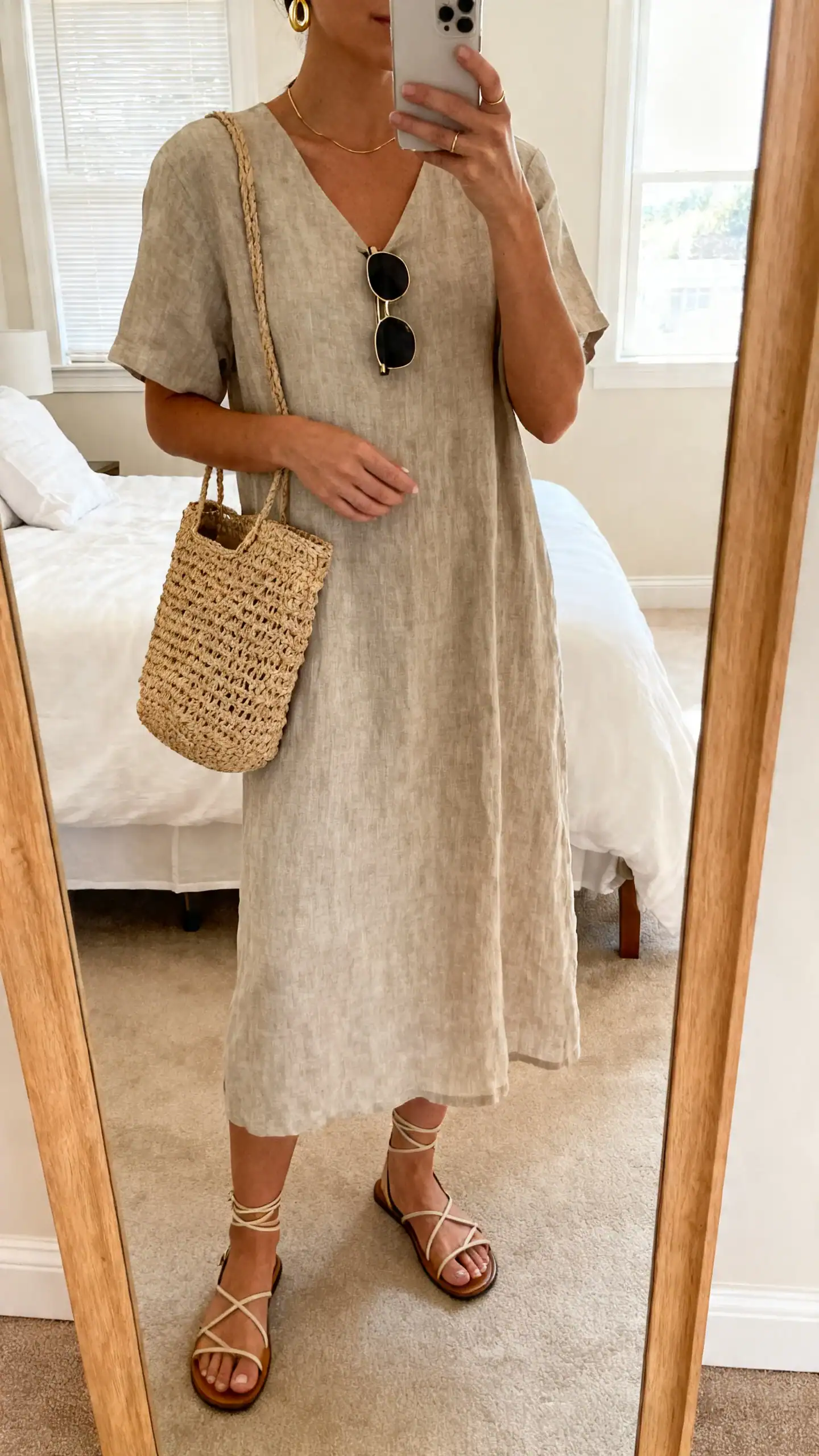 iPhone mirror selfie of a woman wearing a linen midi dress with strappy flat sandals, raffia tote, slim sunglasses, delicate gold hoops, face not visible, cropped at neck, bedroom mirror, morning window light, iPhone photo quality