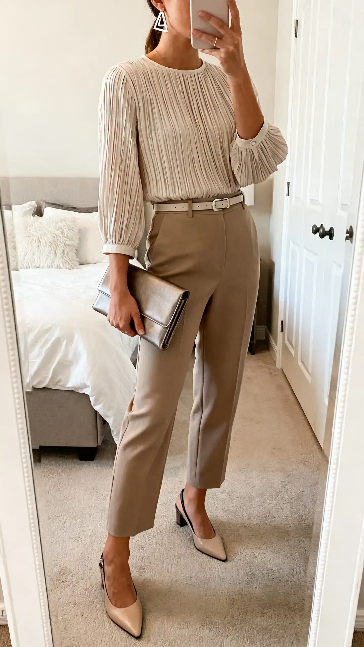 iPhone mirror selfie of a woman wearing a micro-pleated blouse with bracelet-length sleeves, tailored culottes, slingback flats, a minimal belt, geometric earrings, and a sleek clutch, face not visible, side angle, bedroom mirror, soft indoor light, iPhone photo quality.