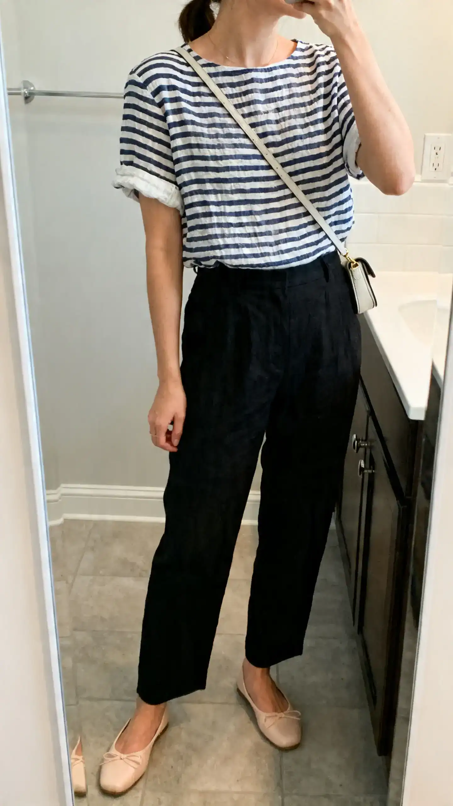 iPhone mirror selfie of a woman wearing a navy-and-white striped linen tee with slightly cuffed sleeves, high-waisted black trousers, ballet flats, a small crossbody bag, face not visible, side angle, bathroom mirror, soft indoor lighting, iPhone photo quality
