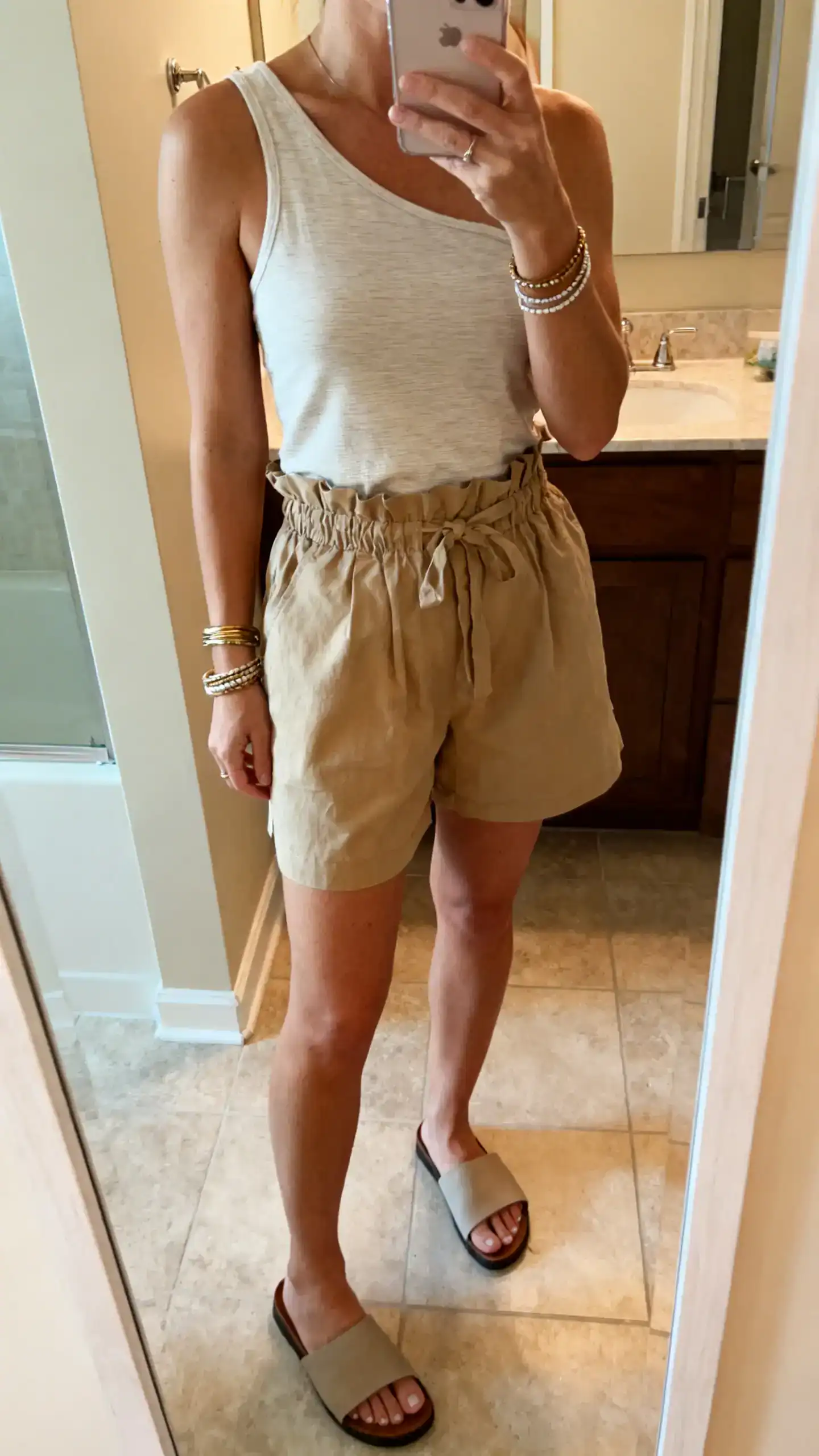 iPhone mirror selfie of a woman wearing a one-shoulder tank with paperbag shorts, slide sandals, and layered bangles, face not visible, cropped at neck, bathroom mirror, warm indoor light, iPhone photo quality.