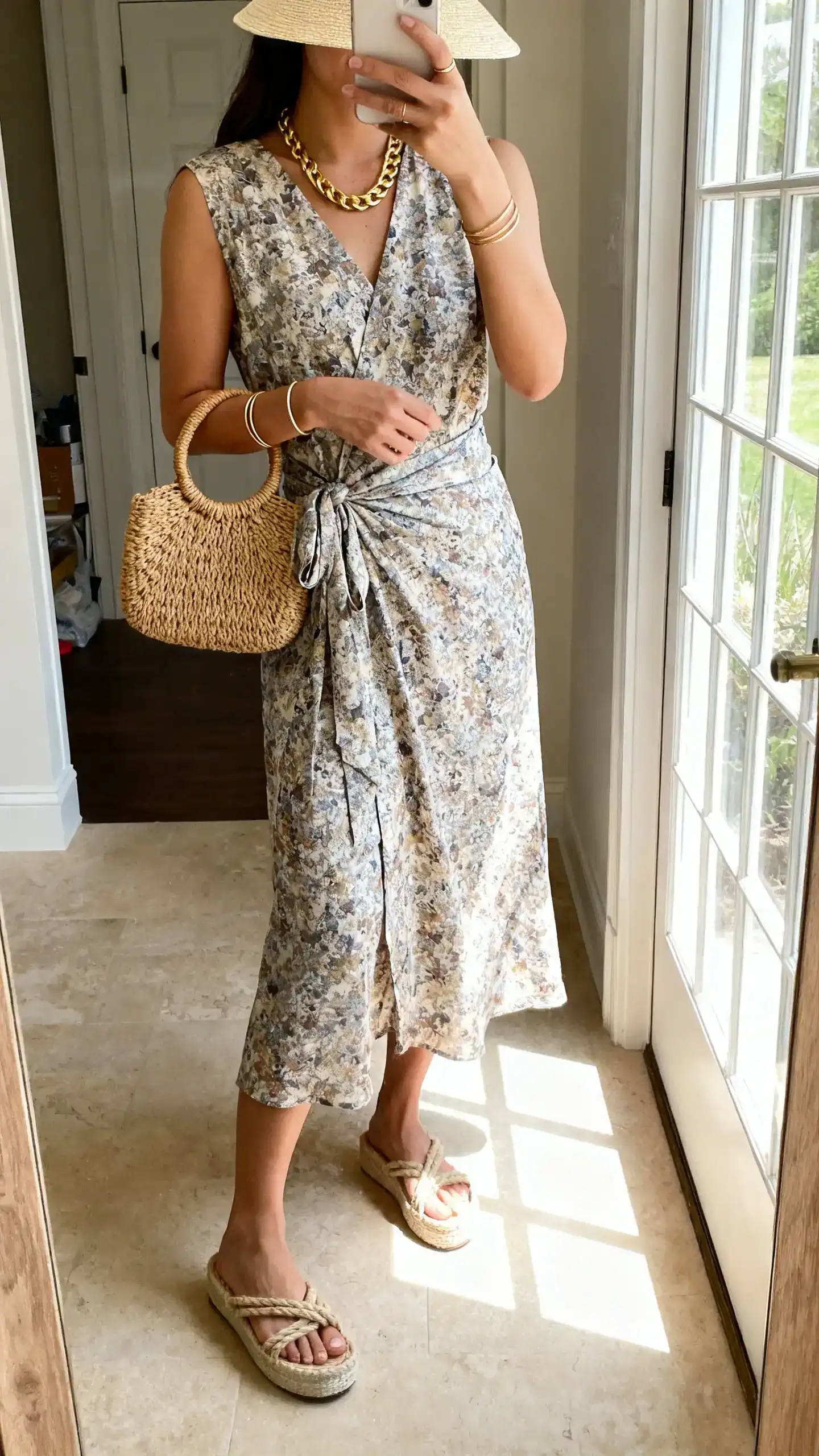 iPhone mirror selfie of a woman wearing a printed midi wrap dress, rope sandals with slight platform, a wicker top-handle bag, a gold chain necklace, slim bangles, and a light straw visor, face not visible, cropped at neck, entryway mirror, natural window light, iPhone photo quality.