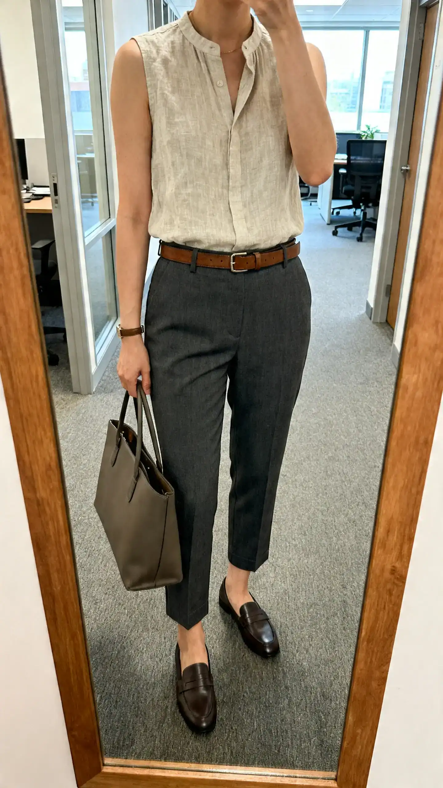iPhone mirror selfie of a woman wearing a sleeveless linen shell tucked into tailored charcoal ankle pants, slim leather belt, loafers, structured tote, face not visible, cropped at neck, office hallway mirror, indoor daylight, iPhone photo quality