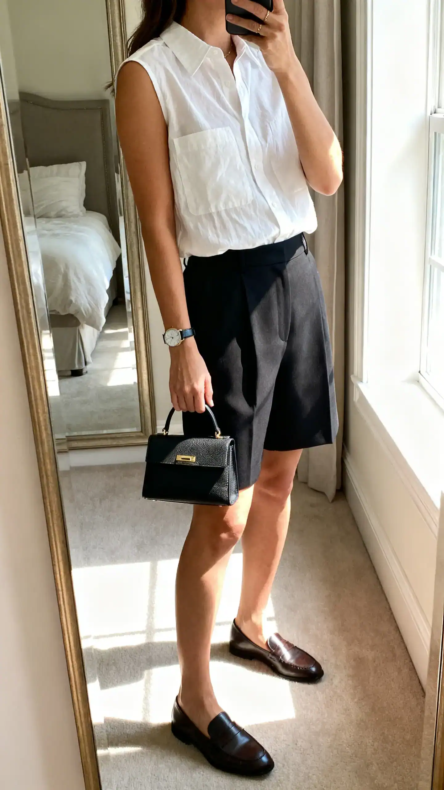 iPhone mirror selfie of a woman wearing a sleeveless white poplin shirt, tailored black shorts, loafers, slim watch, structured mini tote, face not visible, side angle, bedroom mirror, bright morning window light, iPhone photo quality