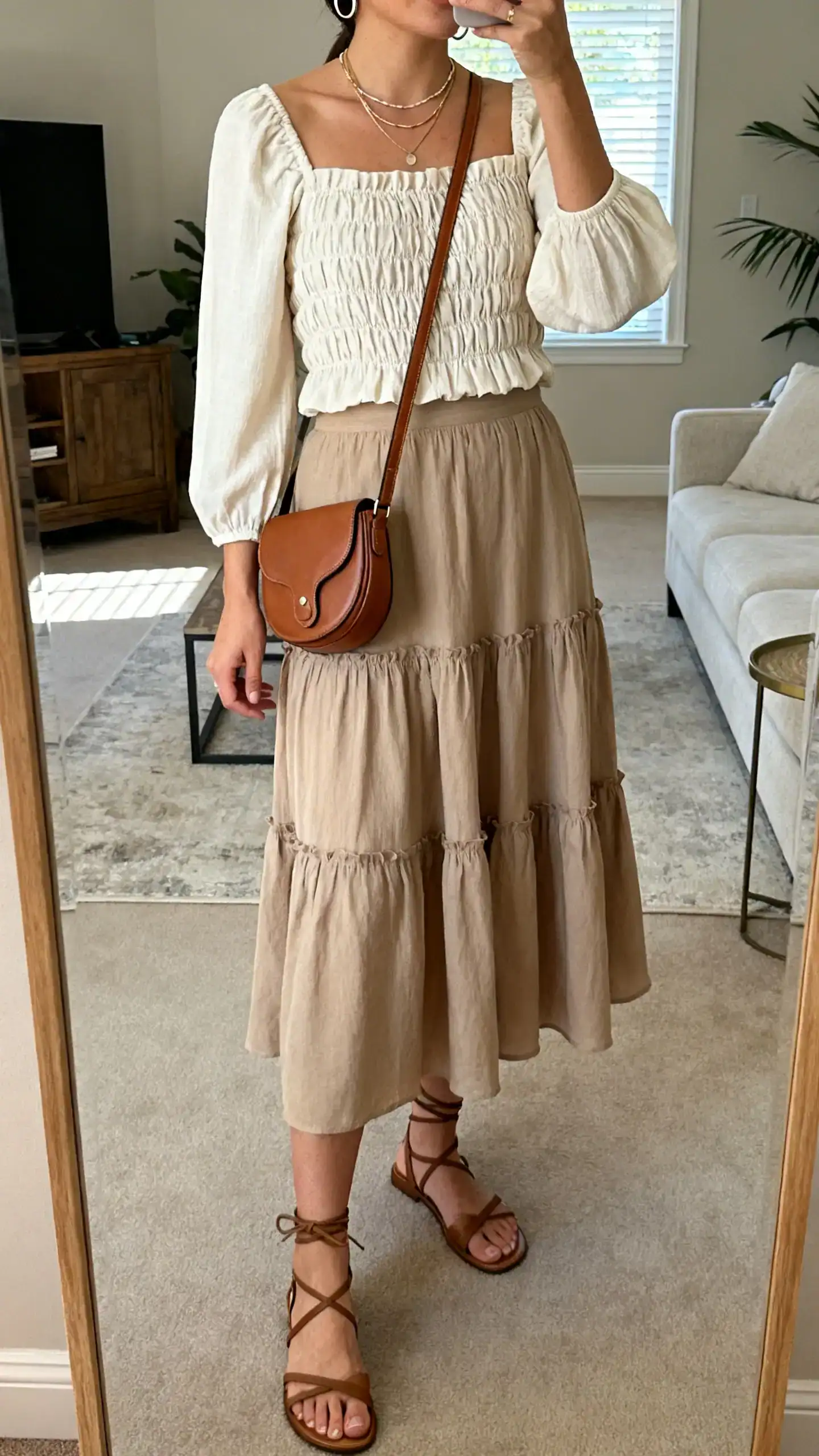 iPhone mirror selfie of a woman wearing a smocked-bodice blouse with elbow-length sleeves, a tiered midi skirt, ankle-strap sandals, a crossbody saddle bag, delicate layered necklaces and slim hoops, face not visible, cropped at shoulders, living-room full-length mirror, natural daylight, iPhone photo quality.