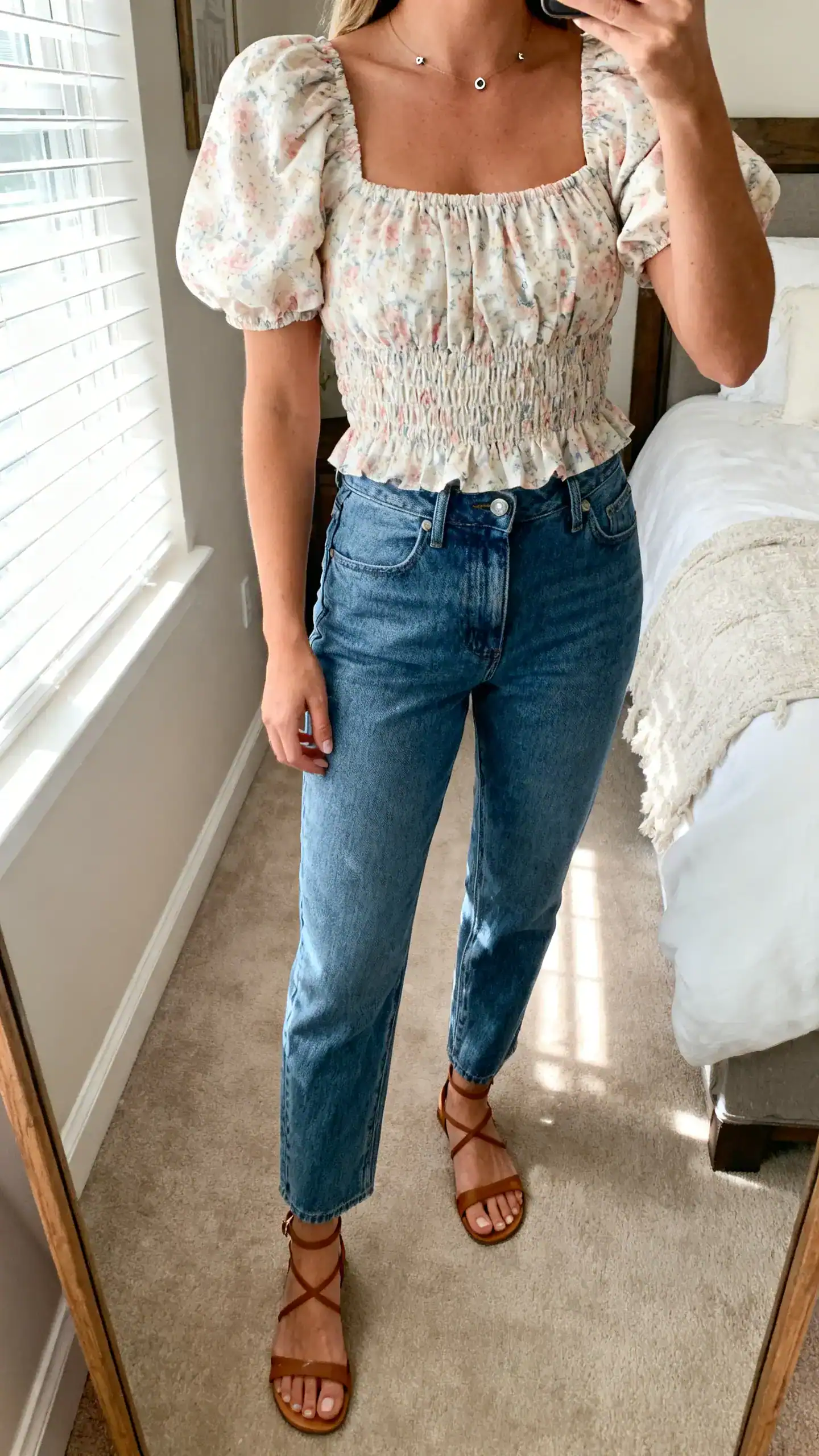 iPhone mirror selfie of a woman wearing a smocked bodice top with puff sleeves in soft floral print, high-waisted straight jeans, ankle-strap sandals, simple studs, face not visible, cropped at neck, bedroom mirror, gentle morning window light, iPhone photo quality