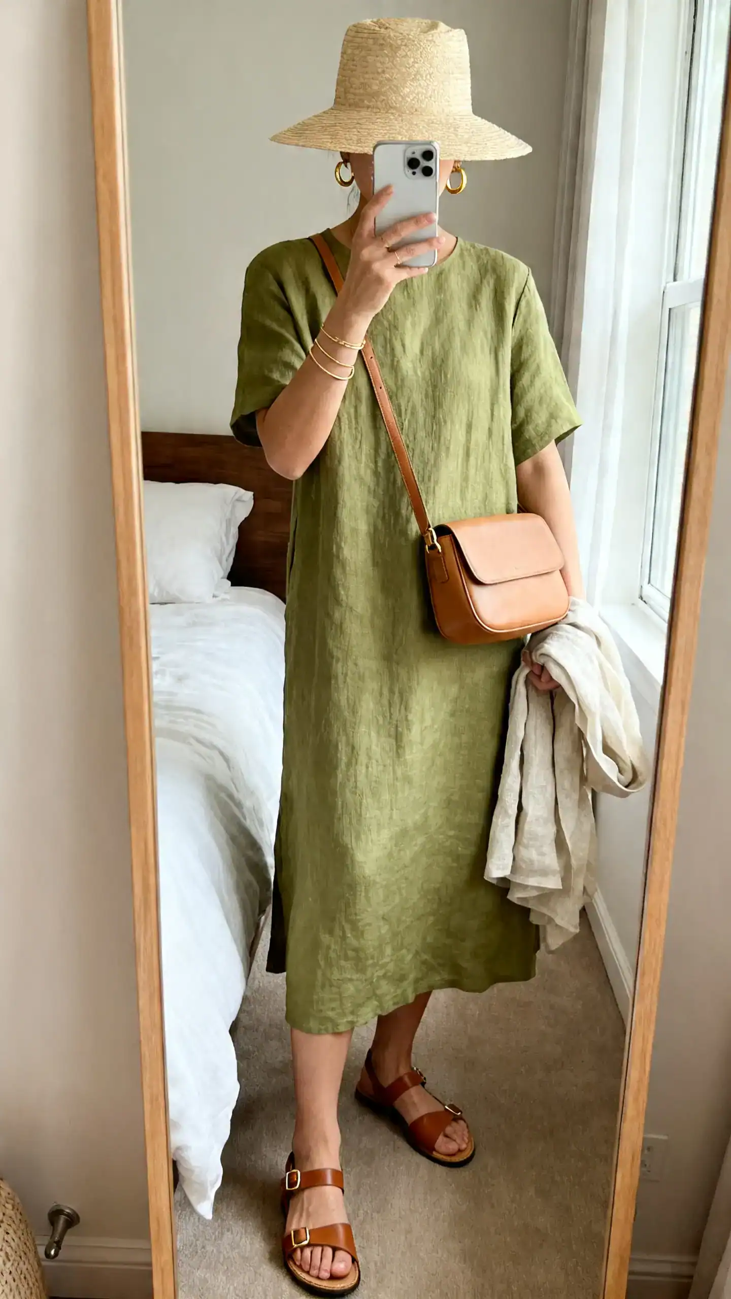 iPhone mirror selfie of a woman wearing a solid linen midi dress in sage, supportive flat leather ankle-strap sandals, a light straw hat, a structured tan crossbody bag, delicate gold hoops and thin bracelets, and a lightweight linen layer over her arm; face not visible, cropped at neck, bedroom mirror, morning window light, iPhone photo quality.