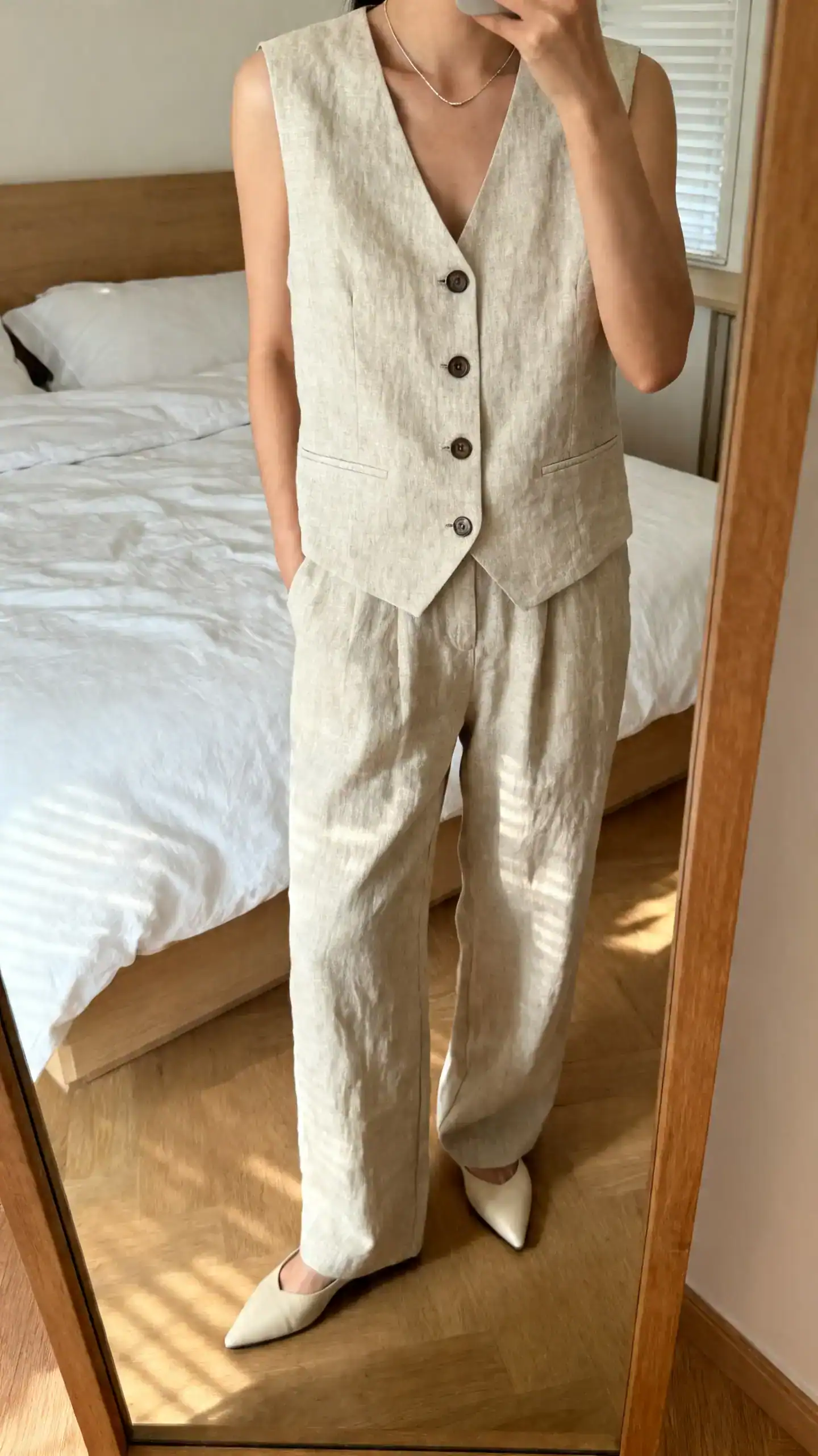 iPhone mirror selfie of a woman wearing a tailored linen vest top buttoned over matching linen trousers, sleek minimal necklace, pointed flats, face not visible, cropped at neck, bedroom mirror, gentle morning light, iPhone photo quality