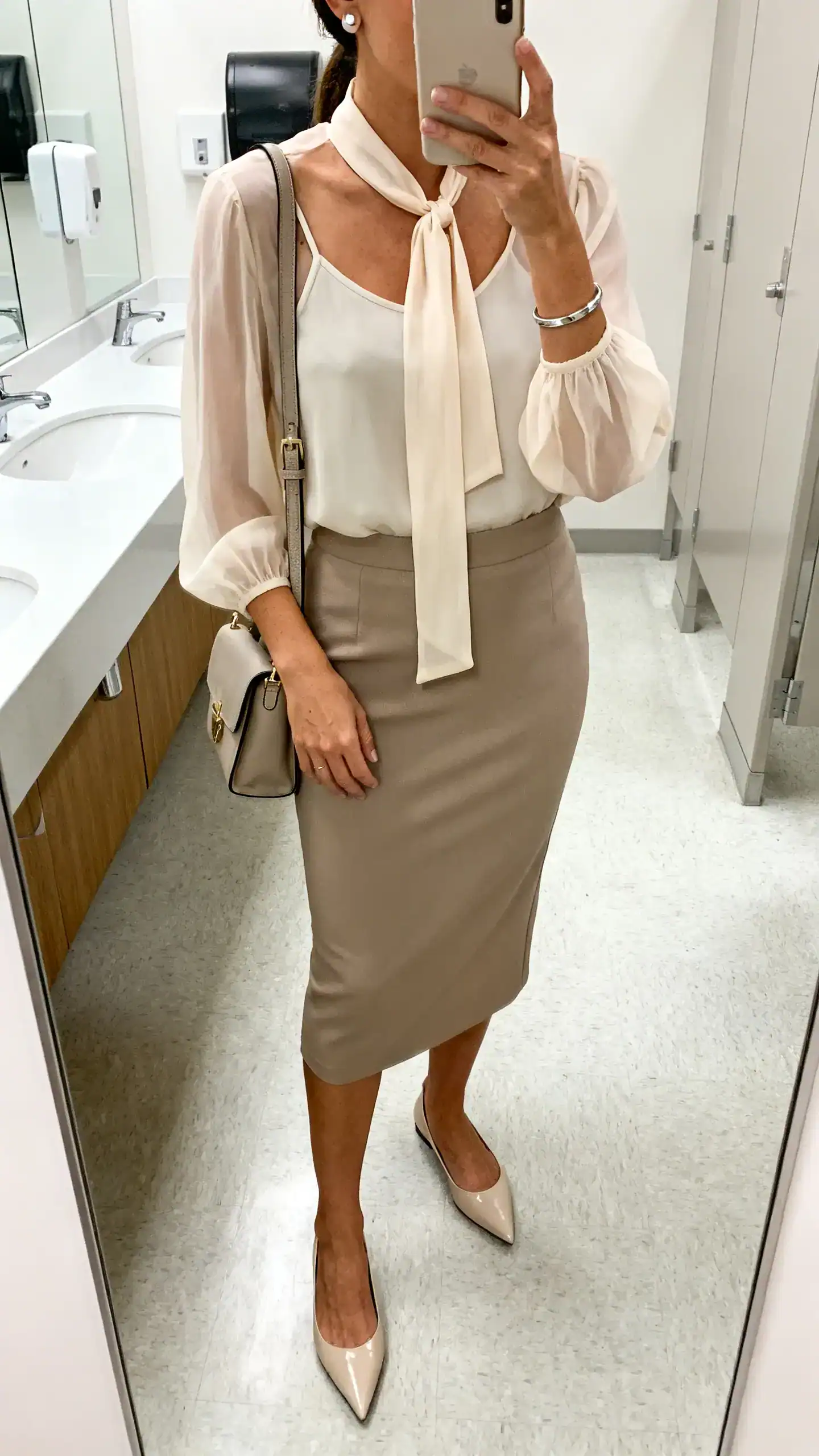 iPhone mirror selfie of a woman wearing a tie-neck blouse in chiffon with long sleeves, a pencil midi skirt, pointed-toe flats, a structured shoulder bag, stud earrings and a sleek bracelet, light camisole underneath, face not visible, cropped at neck, office bathroom mirror, bright indoor lighting, iPhone photo quality.