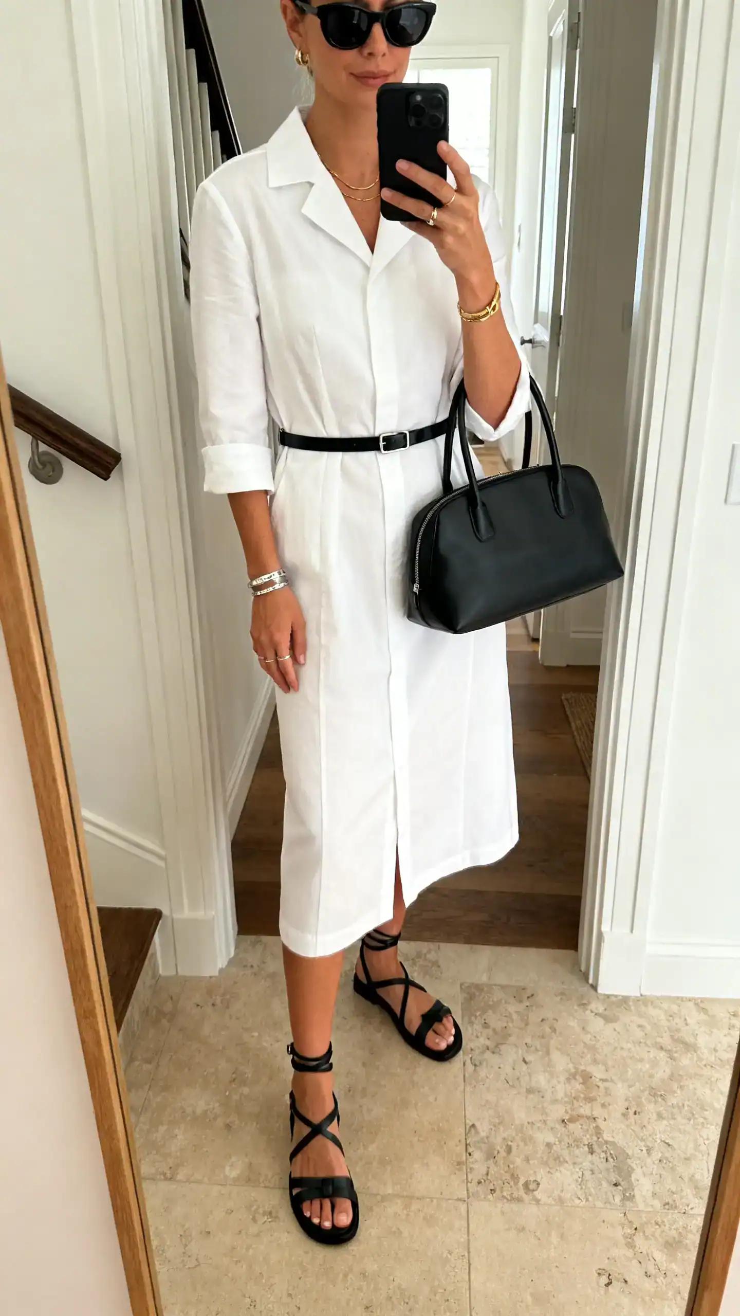iPhone mirror selfie of a woman wearing a white cotton poplin tailored midi dress with black strappy sandals, a thin black belt, structured black top-handle bag, sleek black sunglasses, and minimal silver/gold jewelry; face not visible, cropped at shoulders, entryway mirror, indoor daylight, iPhone photo quality.