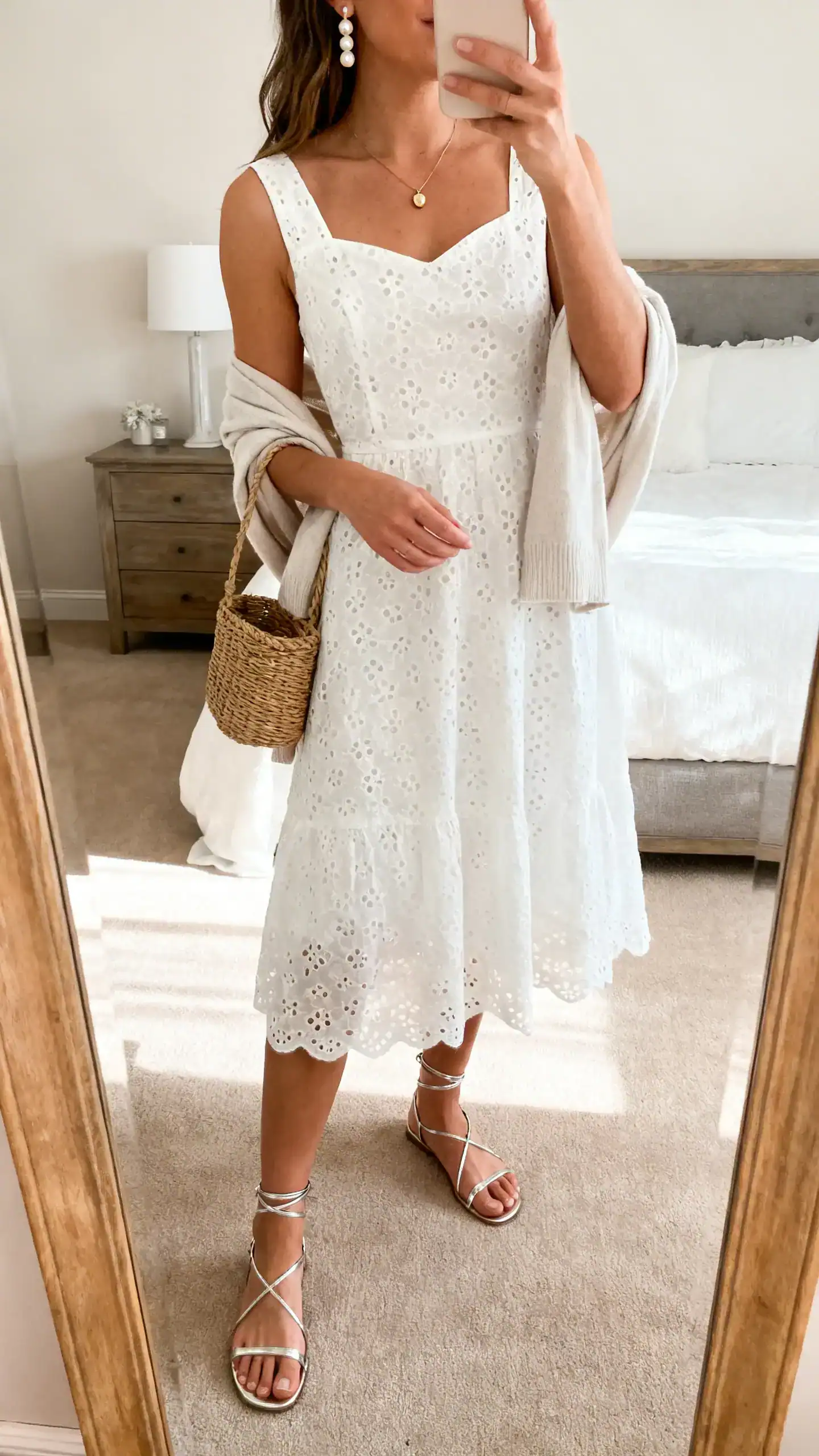 iPhone mirror selfie of a woman wearing a white eyelet midi dress (fit-and-flare) with delicate thin-strap sandals in metallic, a petite pendant necklace, beaded/pearl earrings, a mini basket bag, and a light cardigan draped over shoulders; face not visible, bedroom mirror, soft afternoon light, iPhone photo quality.