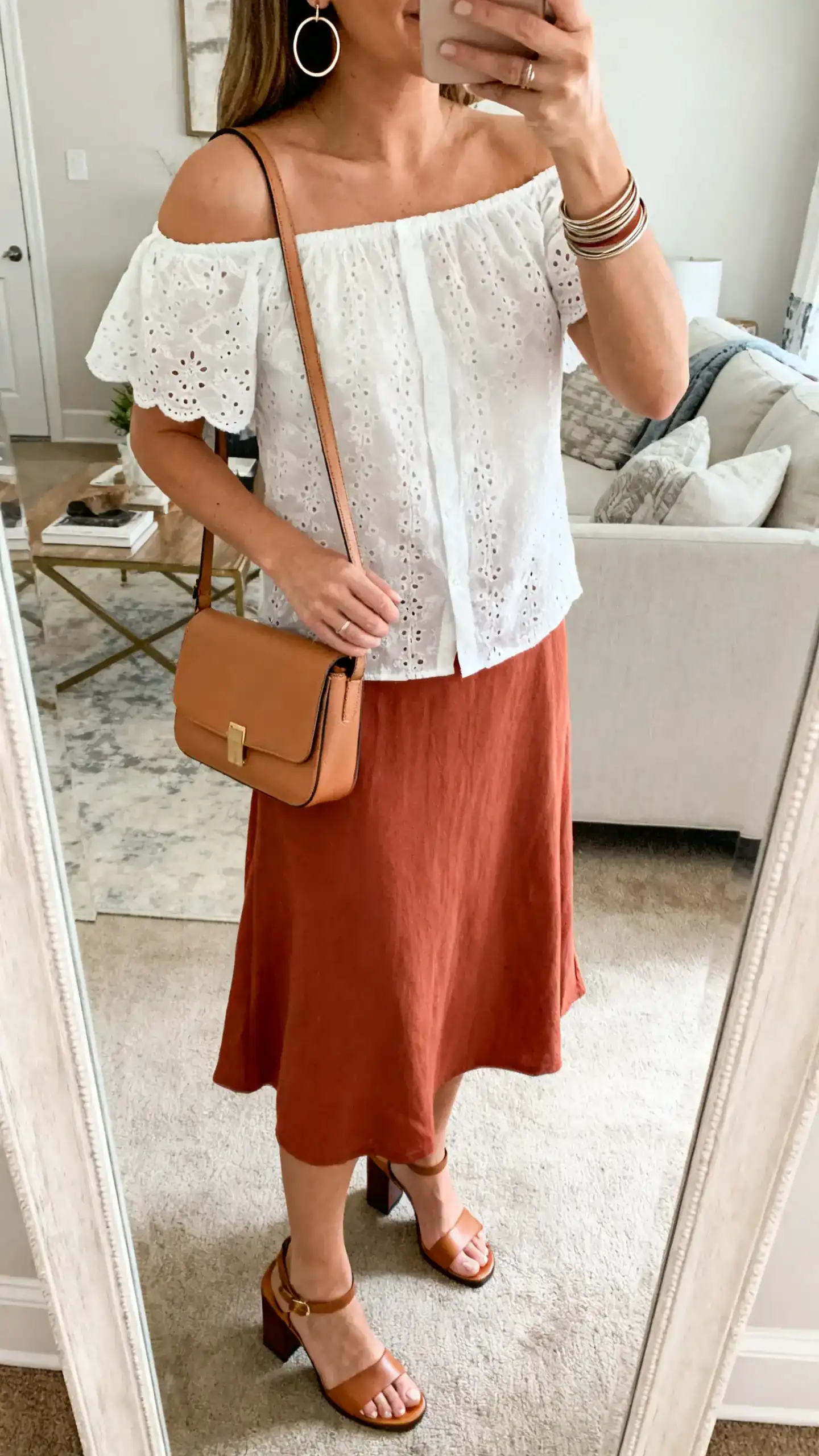 iPhone mirror selfie of a woman wearing a white eyelet off-shoulder blouse with a terracotta midi skirt, block-heel sandals, camel mini shoulder bag, hoop earrings and a stack of thin bangles, face not visible, cropped at shoulders, living-room mirror with natural daylight, iPhone photo quality.