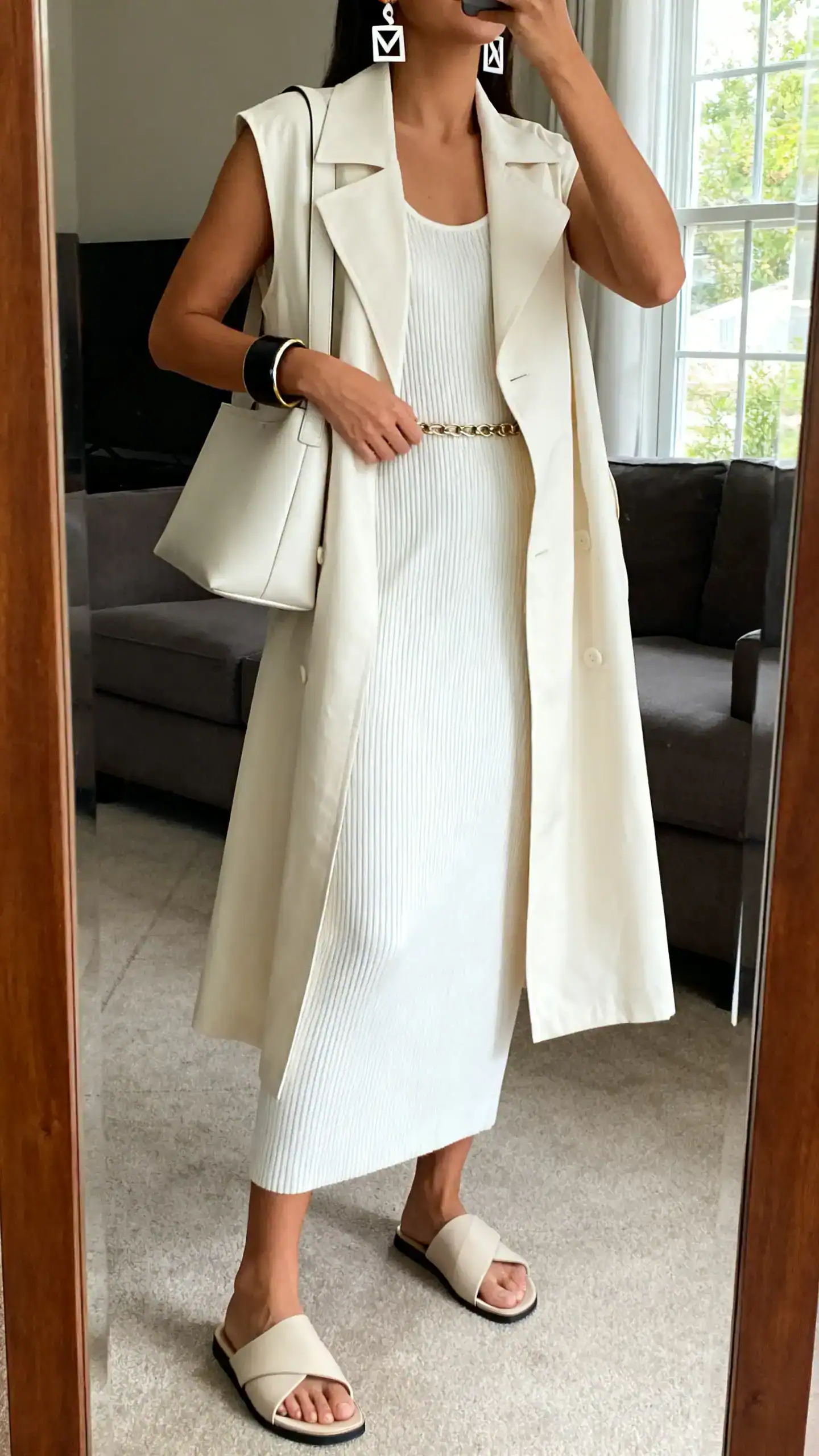 iPhone mirror selfie of a woman wearing a white ribbed knit midi column dress with minimalist leather slides, a cream longline trench vest, sleek shoulder bag, geometric earrings and a bold cuff, optional thin chain belt; face not visible, cropped at neck, living room mirror, natural window light, iPhone photo quality.