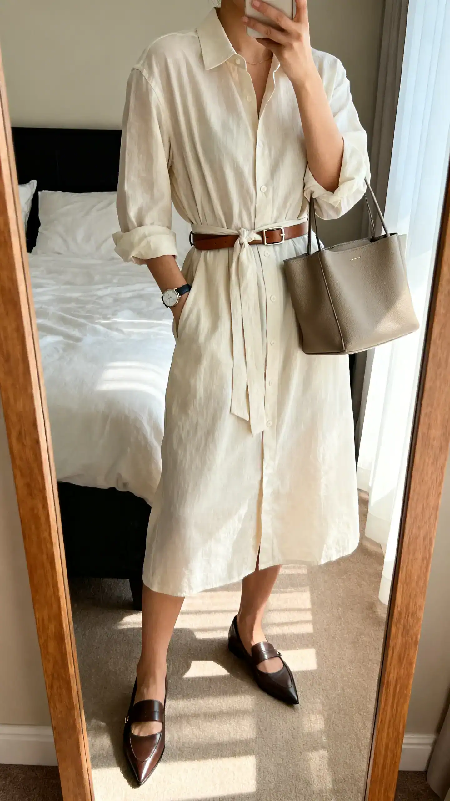 iPhone mirror selfie of a woman wearing an airy cotton-poplin shirt dress with a waist tie, pointed-toe loafers, slim leather belt, structured tote, and a simple watch, face not visible, cropped at neck, bedroom mirror, morning window light, iPhone photo quality