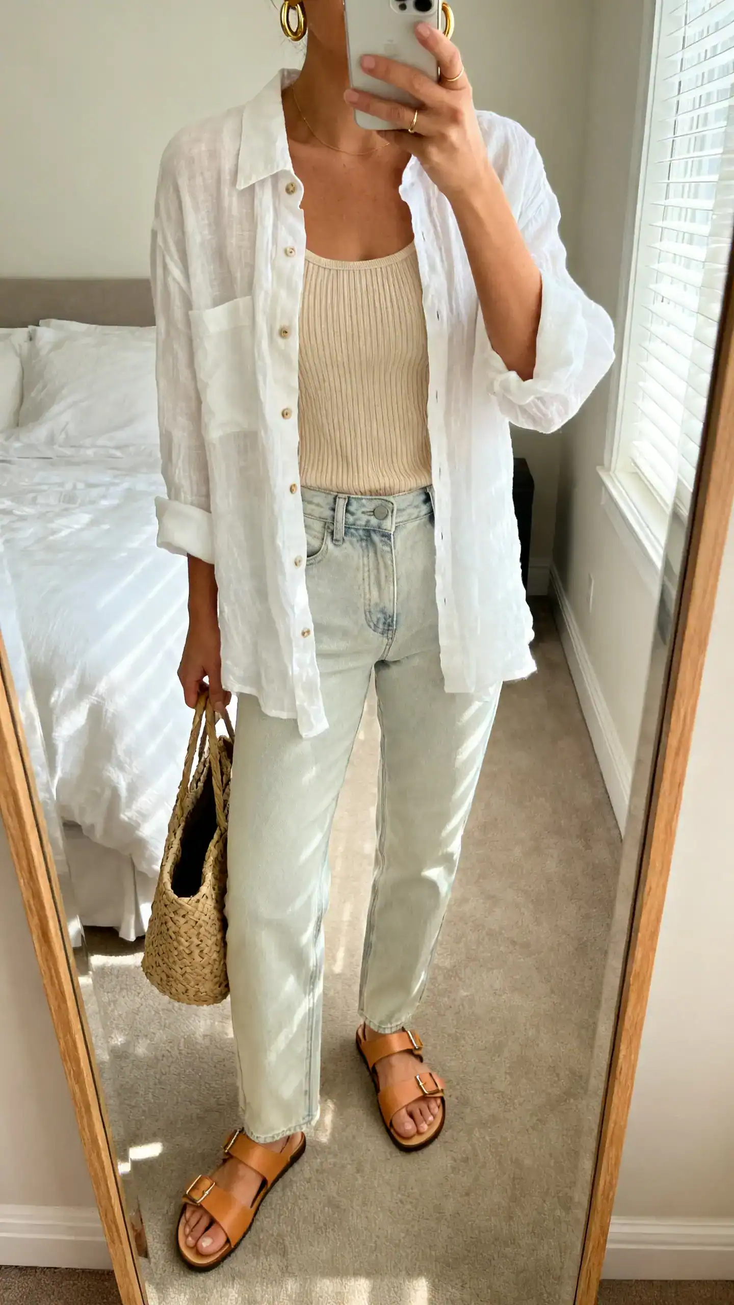 iPhone mirror selfie of a woman wearing an airy white linen button-up left slightly open over a beige ribbed tank, light-wash straight jeans, tan leather sandals, simple gold hoops, woven tote, face not visible, cropped at neck, bedroom mirror, morning window light, iPhone photo quality