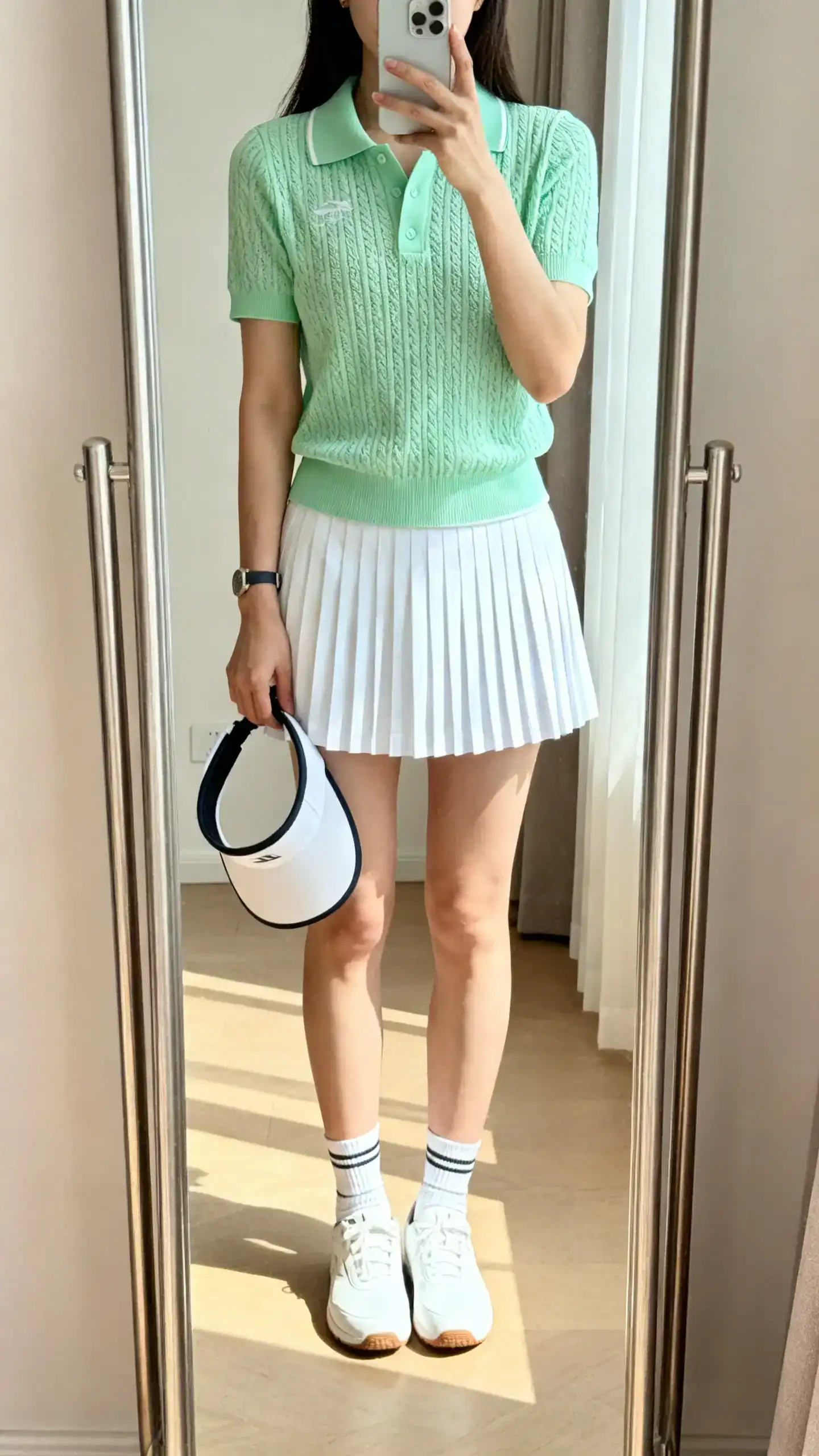 iPhone mirror selfie of a woman wearing an athletic-inspired knit polo in pastel green, pleated tennis skirt in white, low-profile sneakers, sporty socks, visor tucked under arm, face not visible, full-length mirror, bright indoor daylight, iPhone photo quality