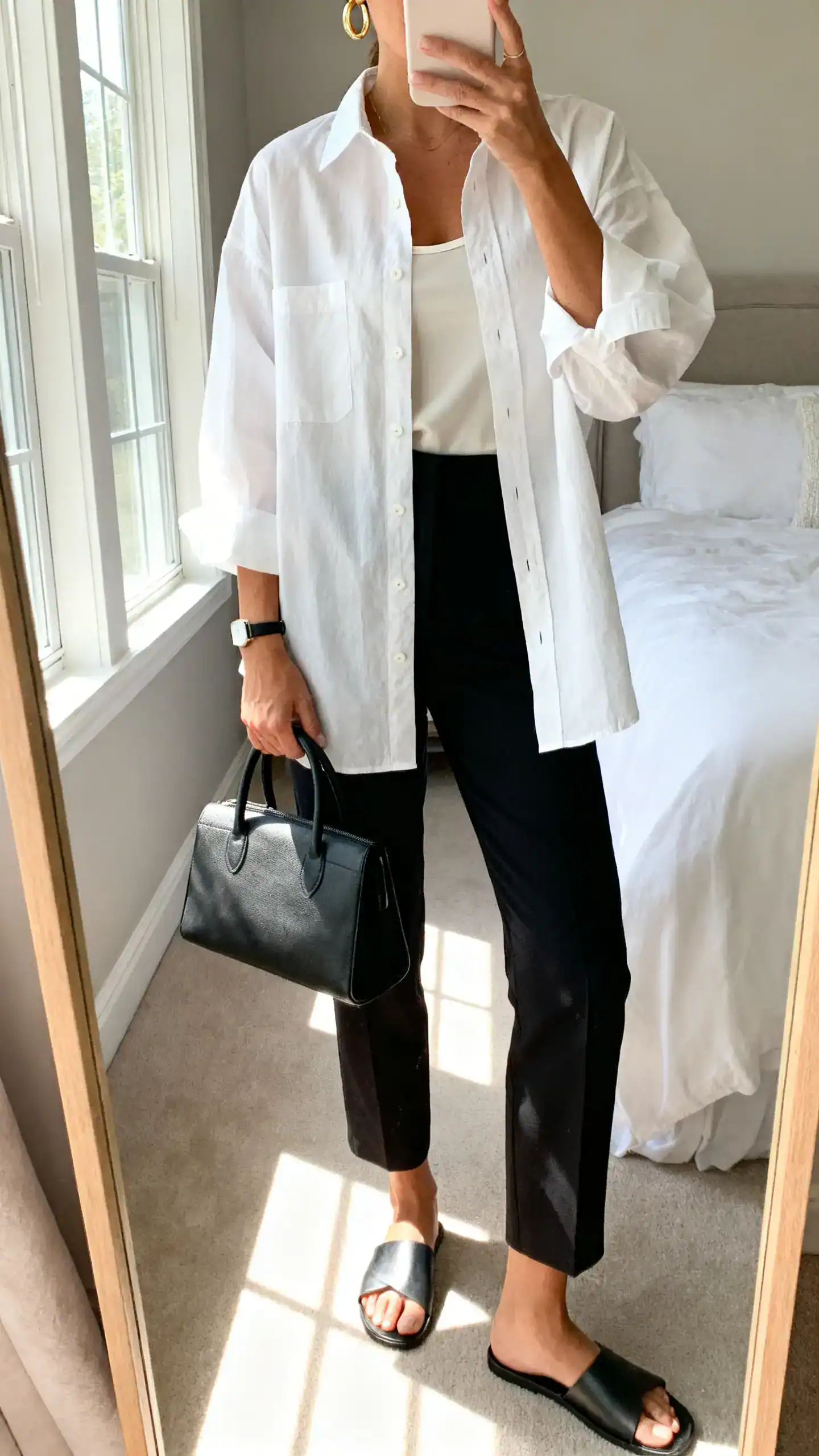 iPhone mirror selfie of a woman wearing an oversized white poplin button-up over a lightweight camisole, tailored ankle trousers in black, minimal leather slides, a structured top-handle bag, simple gold hoops and a slim watch, face not visible, cropped at neck, bedroom mirror, soft morning window light, iPhone photo quality.