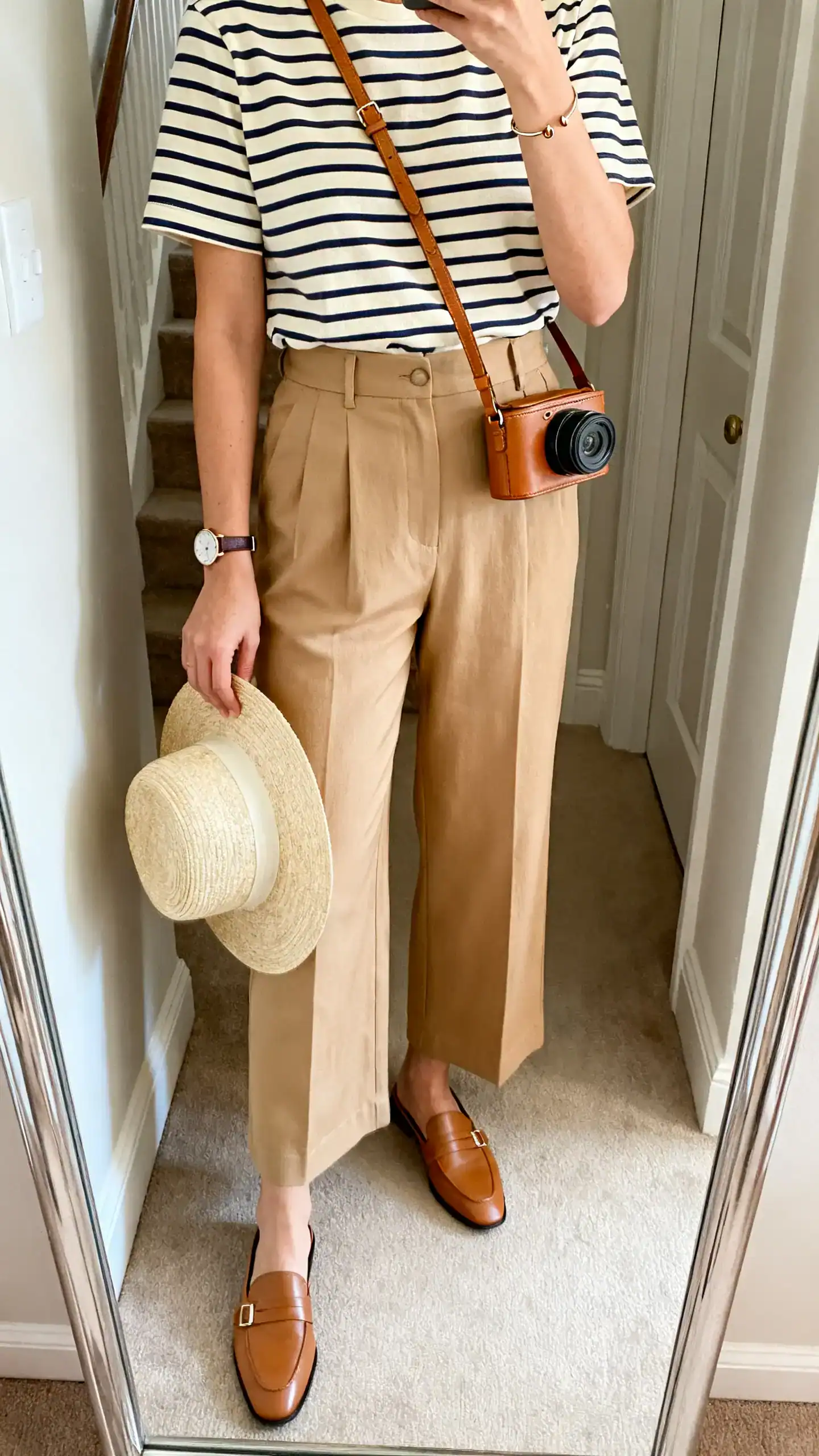 iPhone mirror selfie of a woman wearing high-waisted sand wide-leg trousers, a cream-and-navy Breton striped tee, loafer mules, a tan leather crossbody camera bag, a slim watch with small hoops, and a light straw hat in hand, face not visible, cropped at shoulders, entryway mirror, natural daylight, iPhone photo quality.