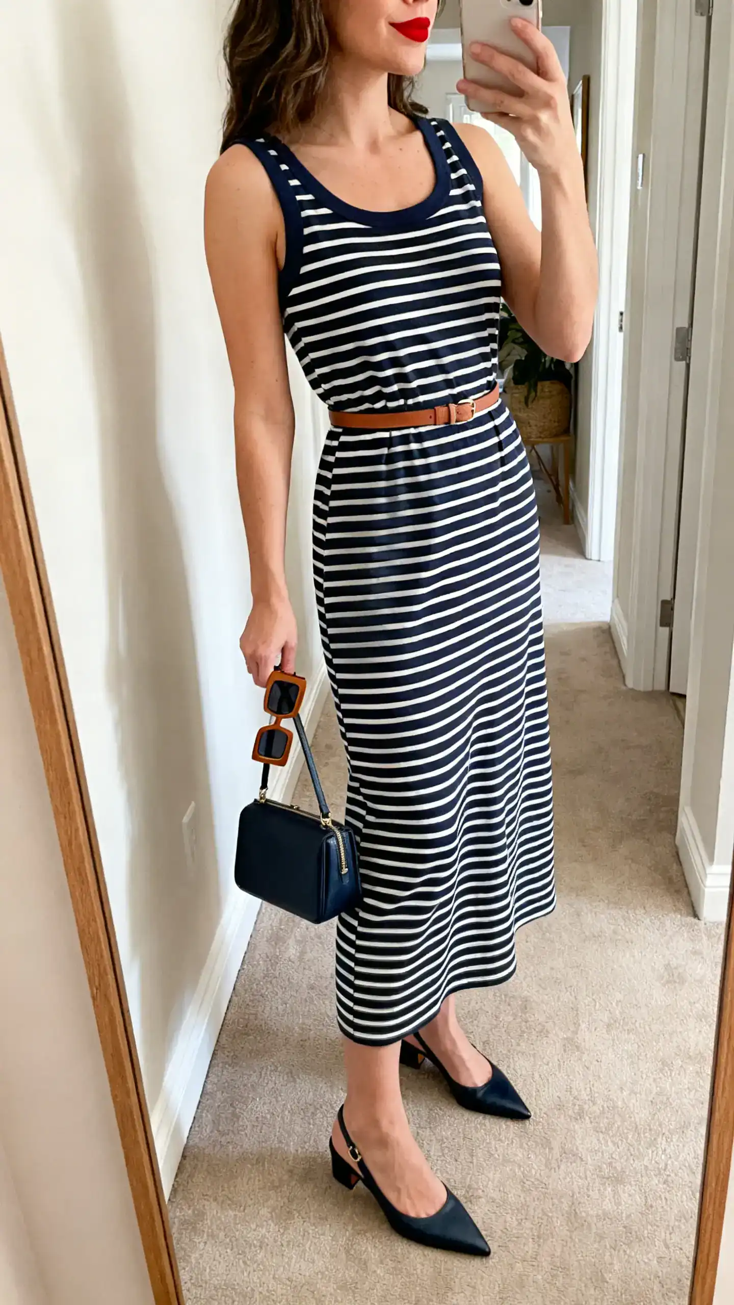 iPhone side-angle mirror selfie of a woman in a boatneck navy-and-white striped midi dress, pointed slingback flats, a structured mini tote, a thin leather belt, square-frame sunglasses in hand, and classic red lipstick visible only on lips, face not visible, hallway mirror, soft morning light, iPhone photo quality.