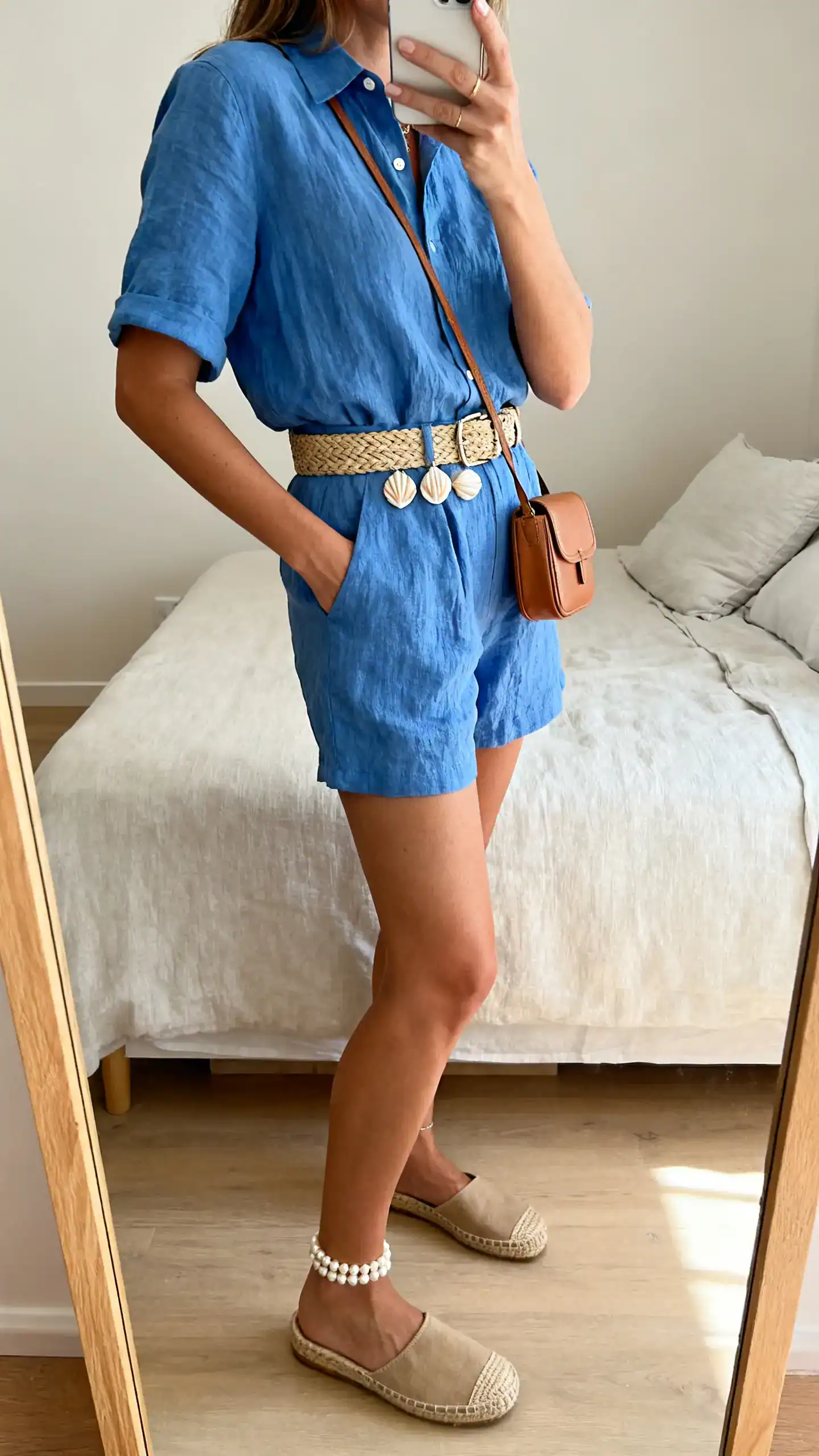 iPhone side-angle mirror selfie of a woman in a Capri-blue linen co-ord (short-sleeve shirt and shorts), flat espadrilles, raffia belt, shell studs with a pearl anklet, compact tan crossbody, face not visible, bedroom mirror, natural daylight, iPhone photo quality.
