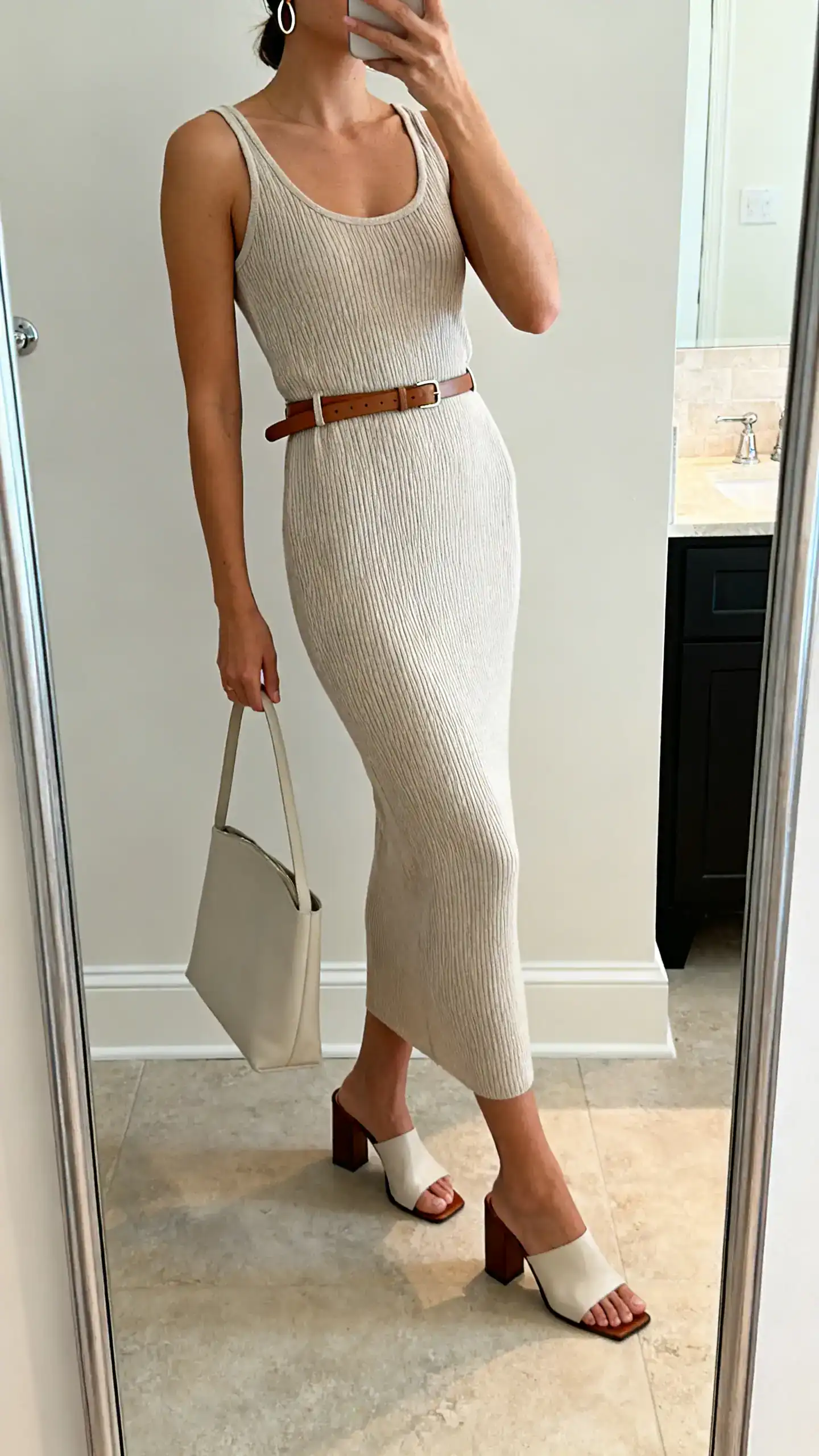 iPhone side-angle mirror selfie of a woman in a ribbed knit tank dress, thin leather waist belt, block-heel sandals, slim hoop earrings, and a minimalist tote, face not visible, cropped at shoulders, bathroom mirror, soft indoor light, iPhone photo quality