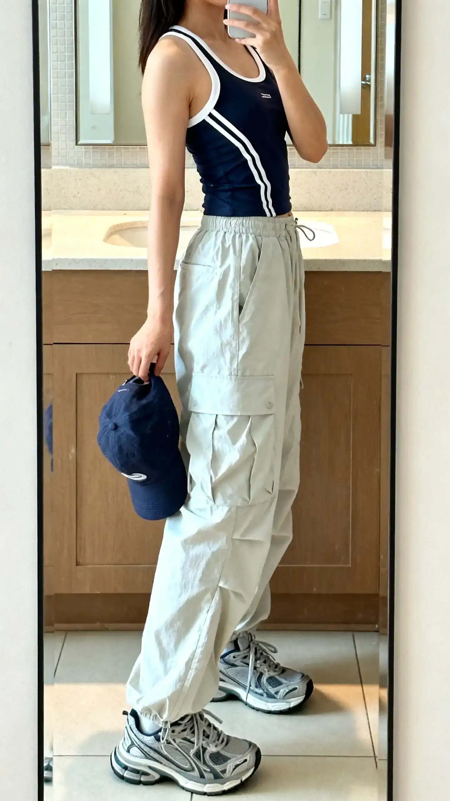 iPhone side-angle mirror selfie of a woman in a sporty-chic racer tank with lightweight parachute pants, chunky sneakers, and a baseball cap in hand, face not visible, bathroom mirror, bright indoor light, iPhone photo quality.