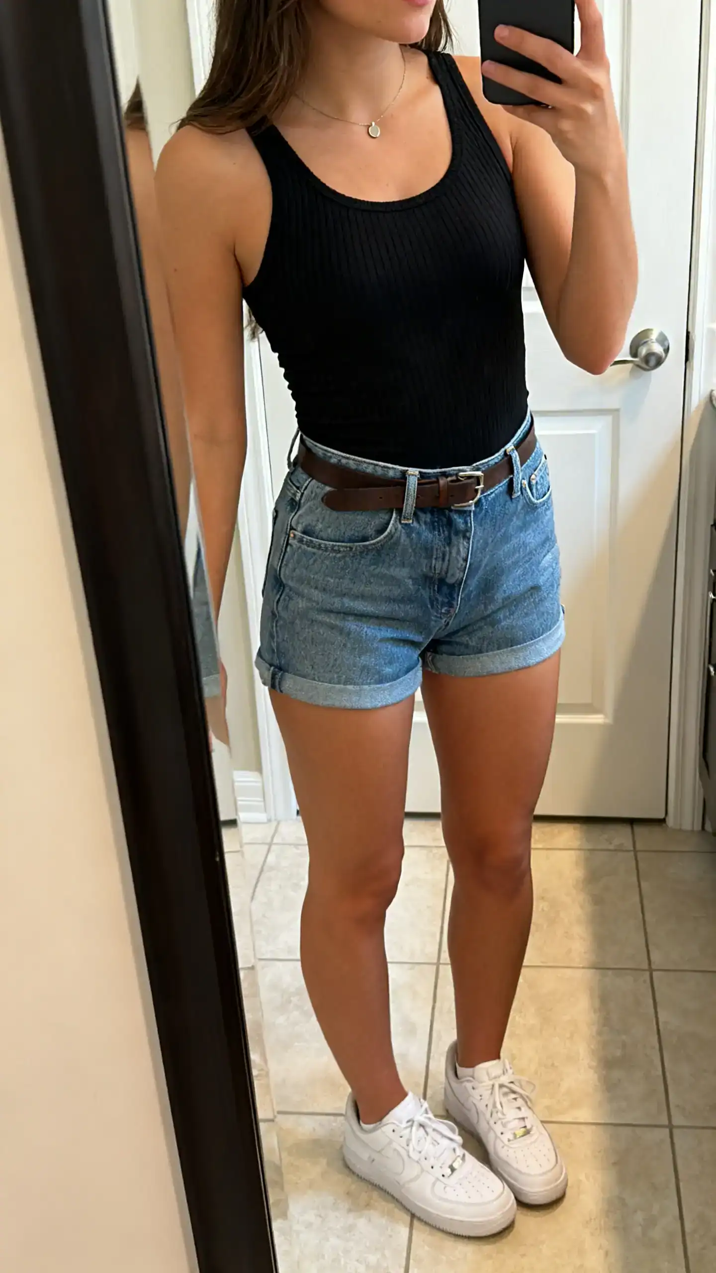 iPhone side-angle mirror selfie of a woman wearing a fitted black ribbed tank, high-waisted denim shorts, white sneakers, thin leather belt, minimal pendant necklace, face not visible, bathroom full-length mirror, soft indoor lighting, iPhone photo quality