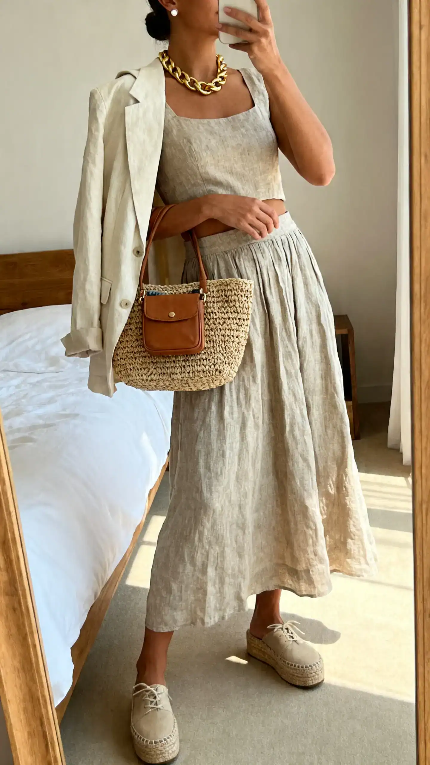 iPhone side-angle mirror selfie of a woman wearing a linen-blend co-ord set (flowy skirt + cropped top), platform espadrilles, a woven tote bag with a mini crossbody tucked inside, chunky gold chain necklace and stud earrings, and a light cotton blazer draped over shoulders; face not visible, cropped at neck, bedroom mirror, daylight, iPhone photo quality.