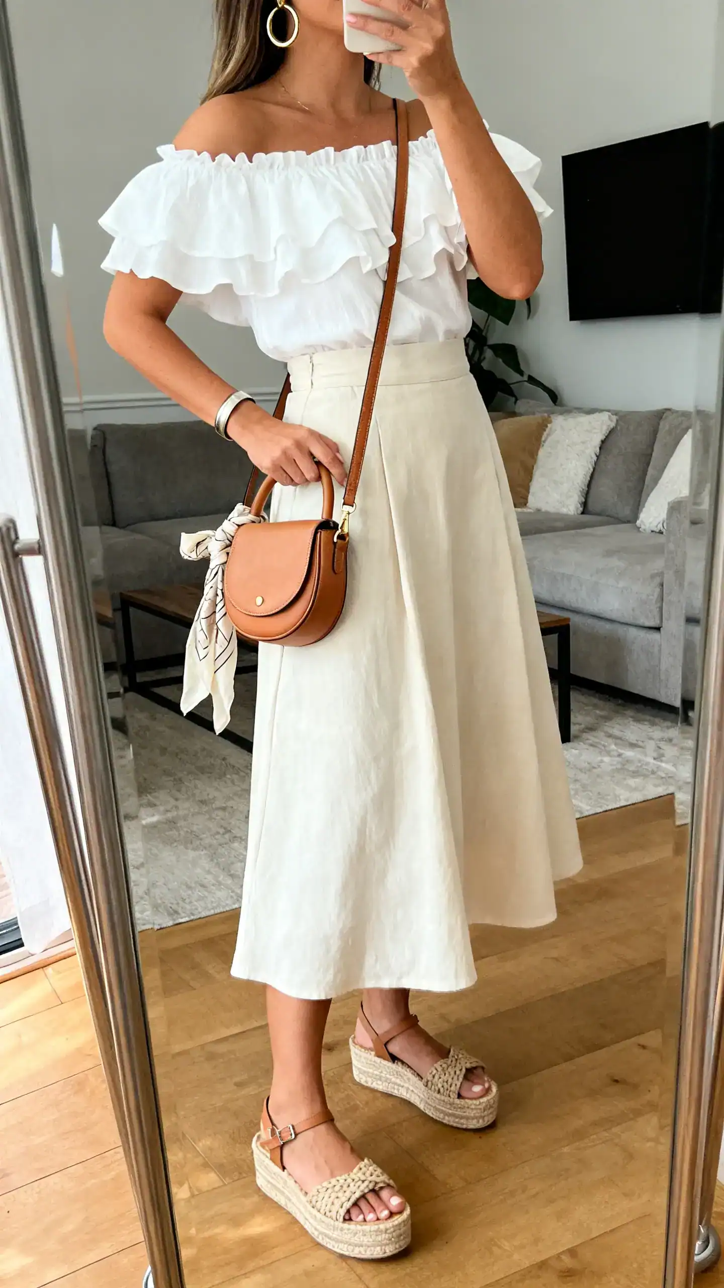 iPhone side-angle mirror selfie of a woman wearing a white off-the-shoulder ruffle top, high-waisted A-line midi skirt in a breezy fabric, woven flatform sandals, tan crossbody saddle bag, statement hoops, simple cuff bracelet, and a lightweight scarf tied to the bag handle; face not visible, living room mirror, natural daylight, iPhone photo quality.