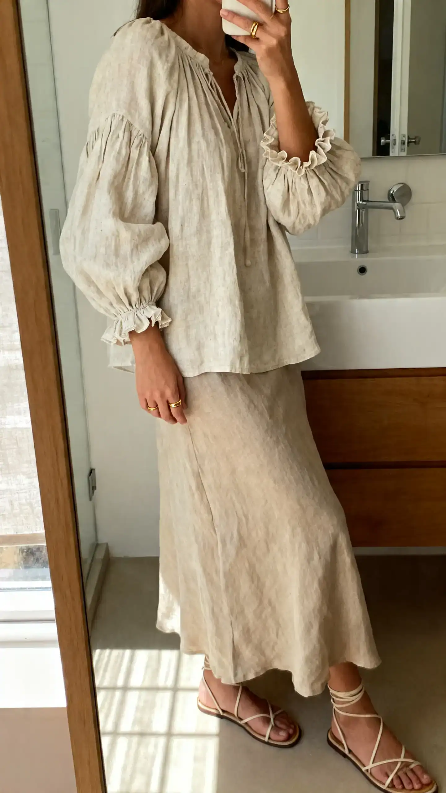 iPhone side-angle mirror selfie of a woman wearing a linen poet blouse with romantic puff sleeves and ruffled cuffs, flowing midi skirt, dainty gold rings, strappy sandals, face not visible, bathroom mirror, soft morning light, iPhone photo quality