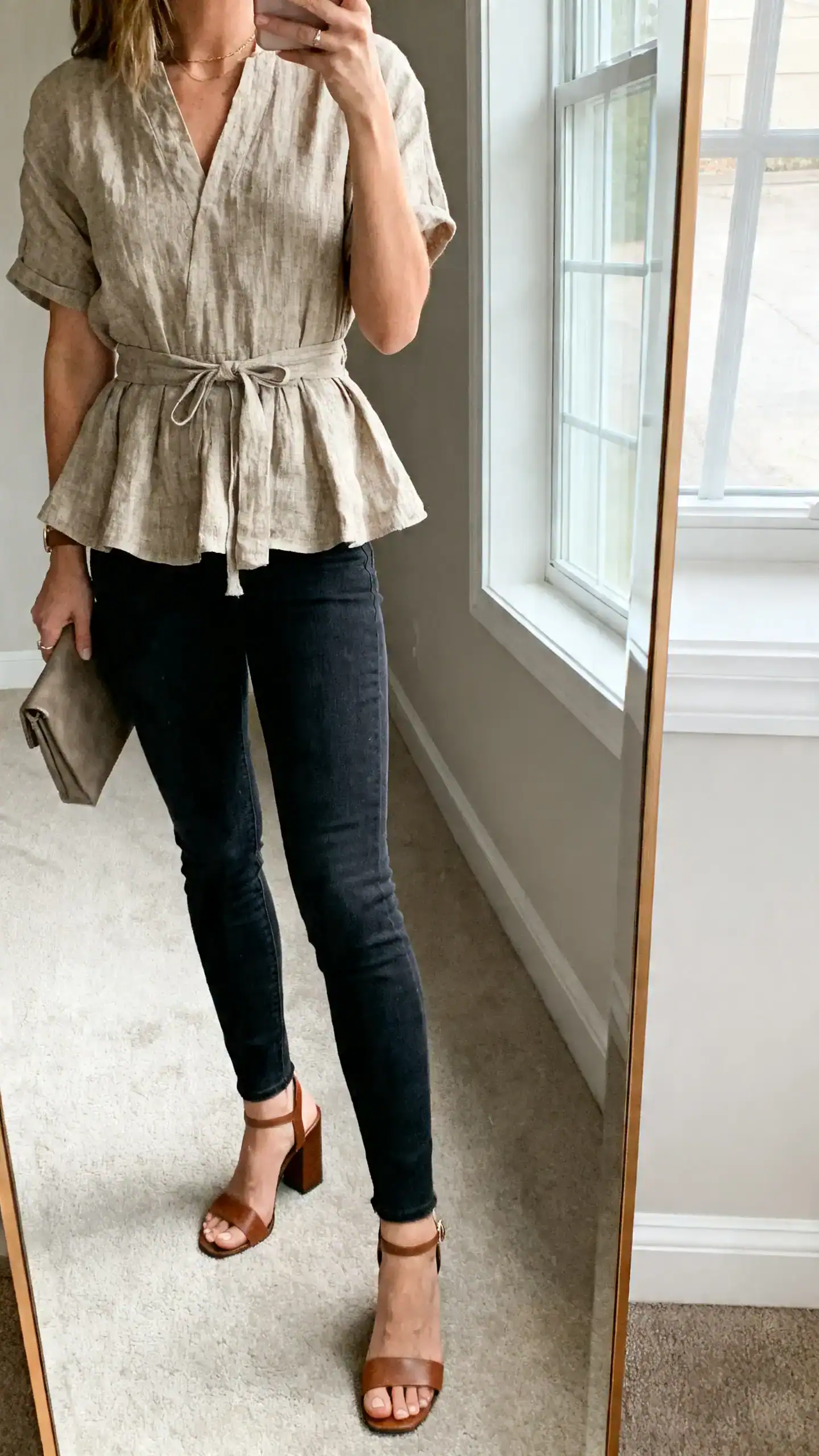 iPhone side-angle mirror selfie of a woman wearing a linen peplum top that cinches at the waist with dark skinny jeans and block-heel sandals, small clutch, face not visible, cropped at shoulders, full-length mirror, soft window light, iPhone photo quality