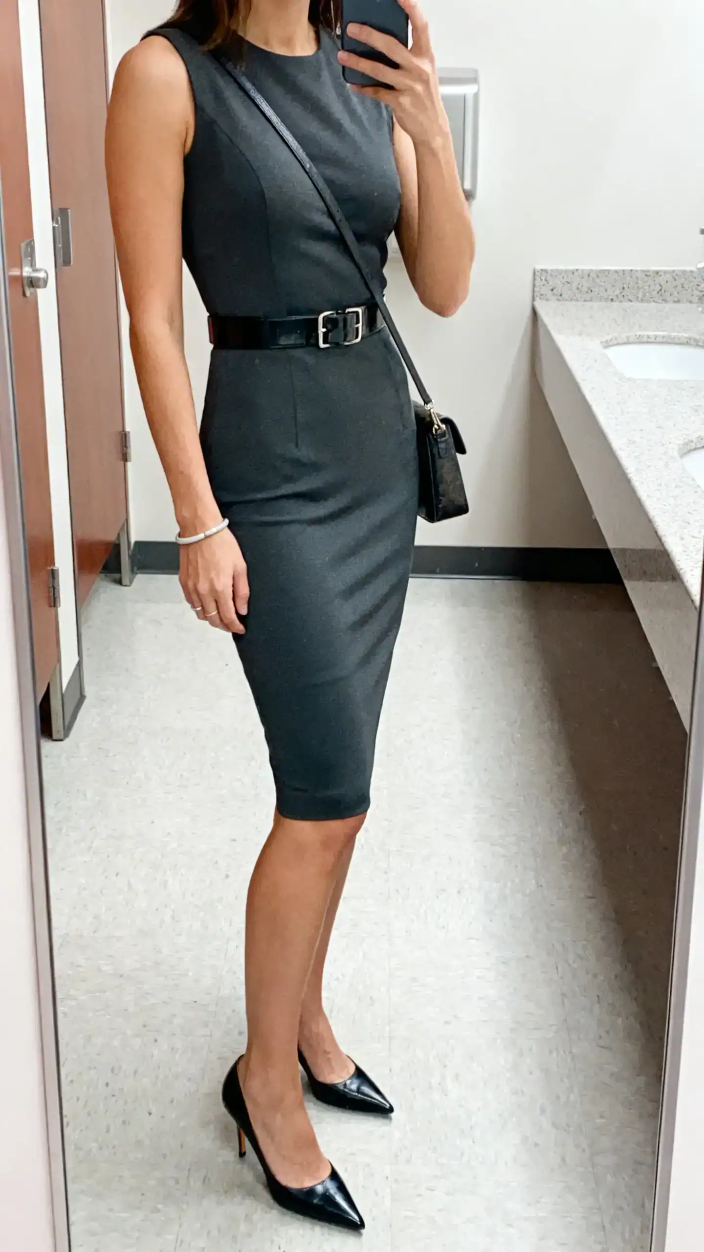 iPhone side-angle selfie of a woman wearing a sleeveless sheath dress with stretch, sleek belt, pointed pumps, simple bracelet, and a structured crossbody, face not visible, cropped at shoulders, office restroom mirror, bright indoor lighting, iPhone photo quality