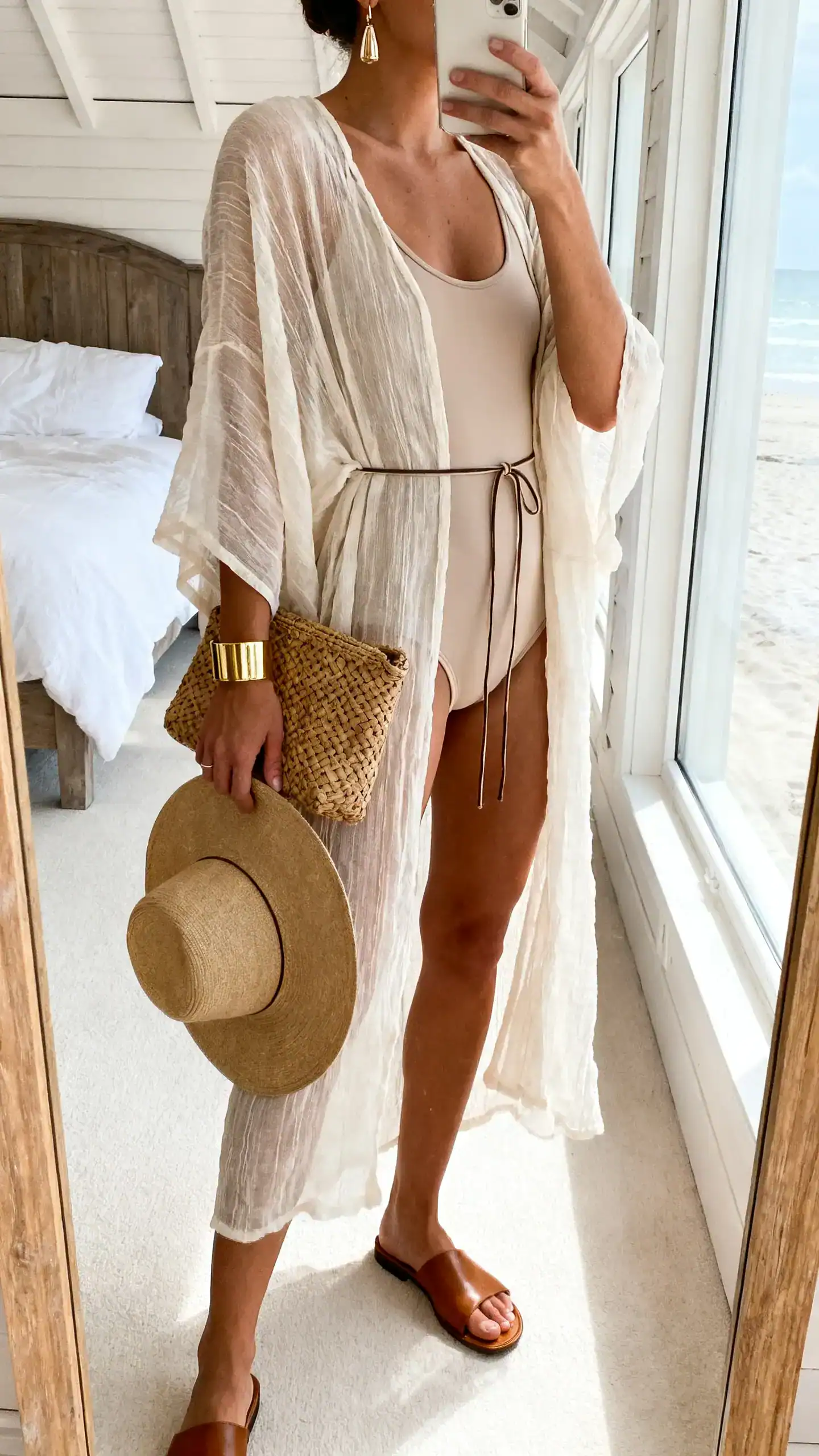 iPhone side-view mirror selfie of a woman in a lightweight gauze kaftan over a chic one-piece cover-up, leather slide sandals, woven clutch, gold cuff and drop earrings, wide-brim hat in hand, thin waist cord tied, face not visible, beach-house bedroom mirror, bright window light, iPhone photo quality.