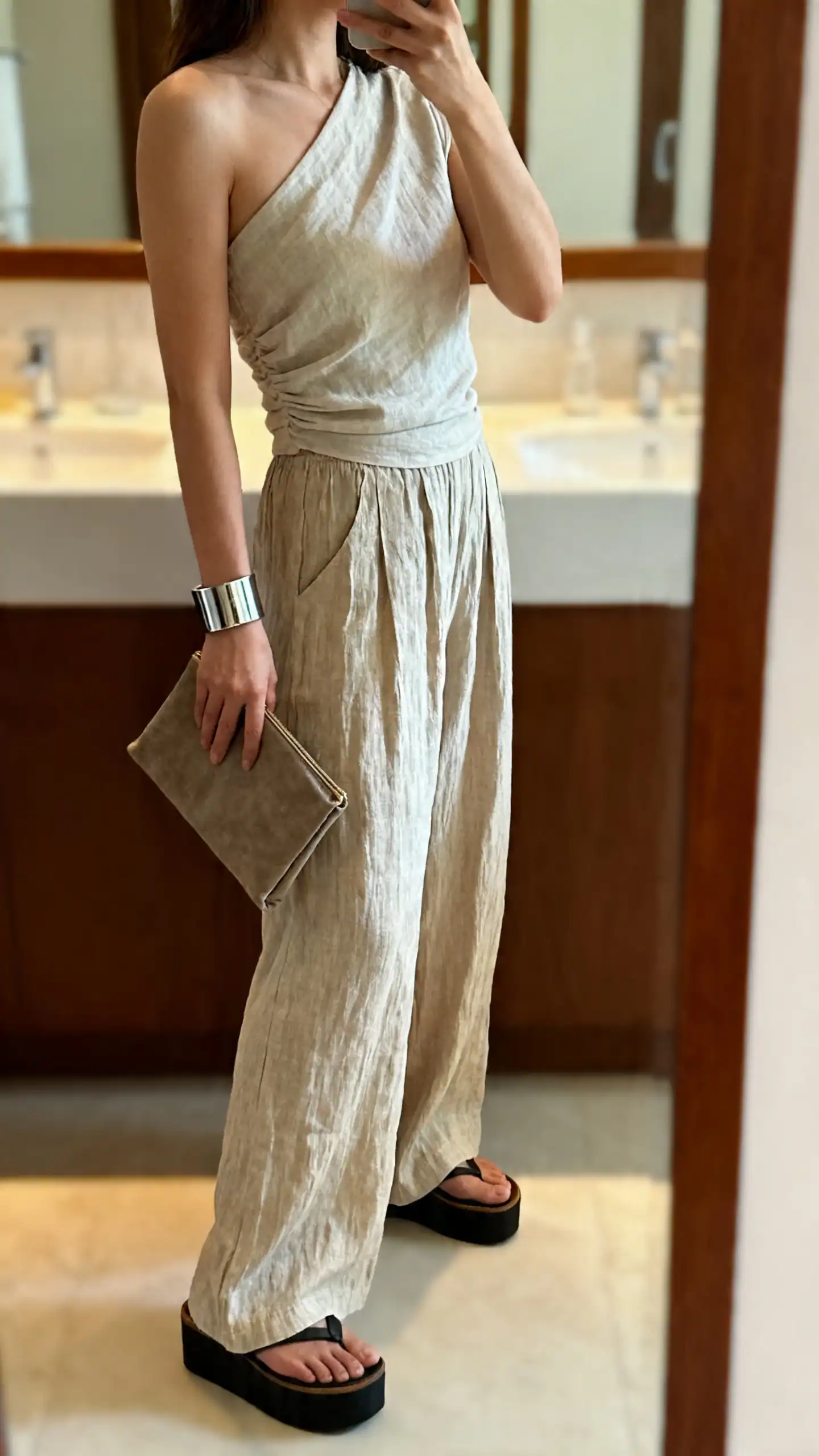 iPhone side-view mirror selfie of a woman in a one-shoulder top with linen palazzo pants, platform slides, cuff bracelet, clutch bag, face not visible, cropped at shoulders, bathroom mirror, warm indoor light, iPhone photo quality