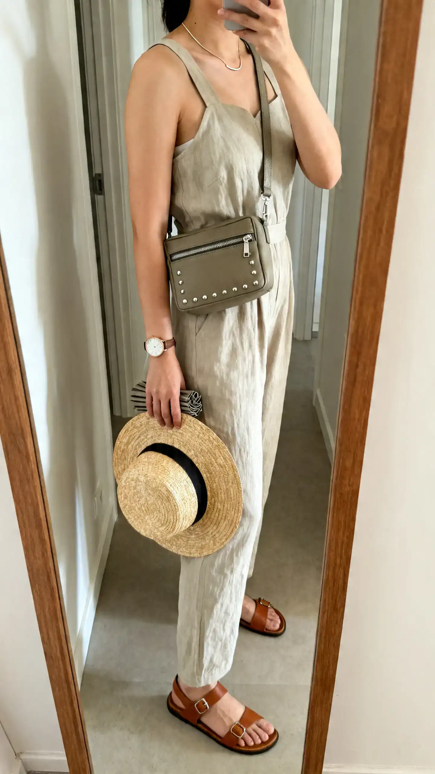 iPhone side-view mirror selfie of a woman wearing a breathable Tencel jumpsuit with wide straps, supportive leather sandals, a medium crossbody with zipper pockets, refined studs, a slim necklace, a watch, and a foldable straw fedora in hand; face not visible, hallway mirror, natural light, iPhone photo quality.