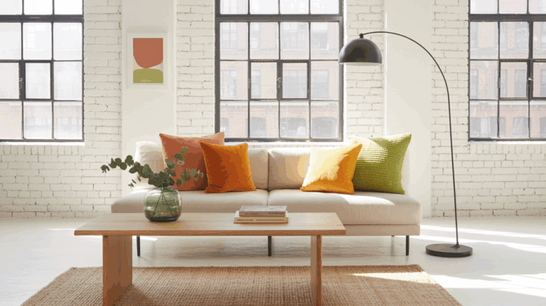 Wide shot, sunlit industrial loft living room with white-painted brick walls and matte black steel-framed windows; a low-profile neutral sofa styled with citrus-colored pillows: tangerine velvet, lemon yellow linen, and lime green boucle; bleached oak coffee table on a natural jute rug; a black arc floor lamp; single abstract print in coral and chartreuse on the wall; jade glass vase with fresh eucalyptus on the coffee table; modern, bright, effortless mood; crisp natural daylight streaming in.