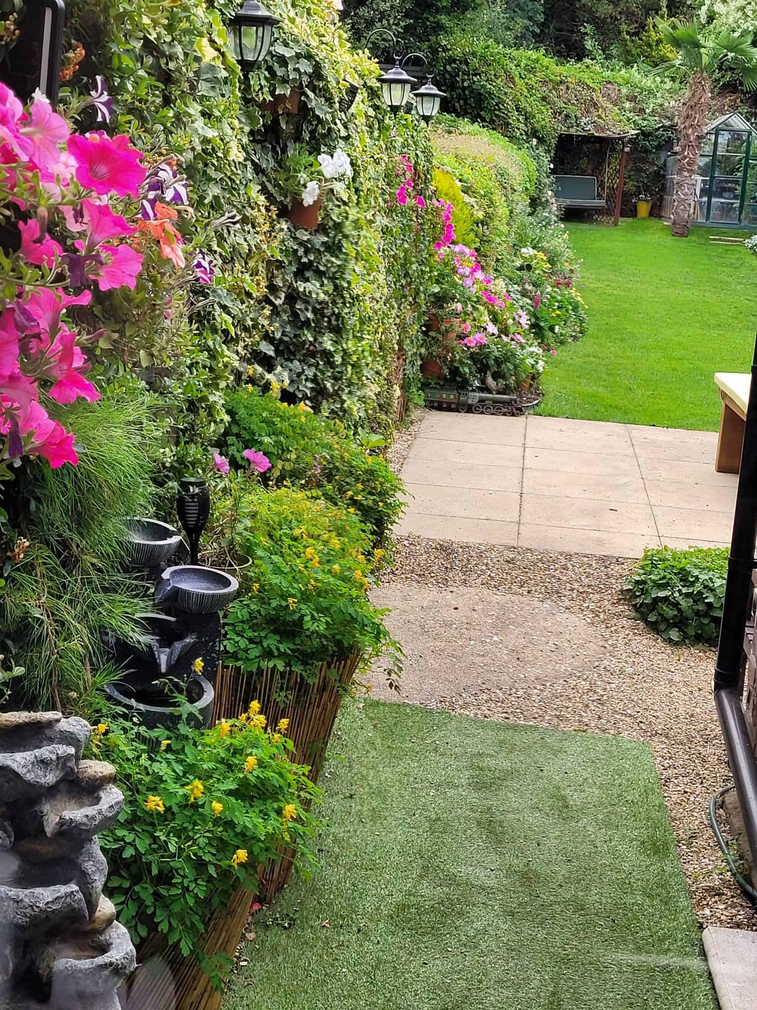Welcoming entrance to Alex’s English cottage garden featuring colourful flower borders, climbing ivy, lanterns, a lush green lawn, and a greenhouse in the background.
