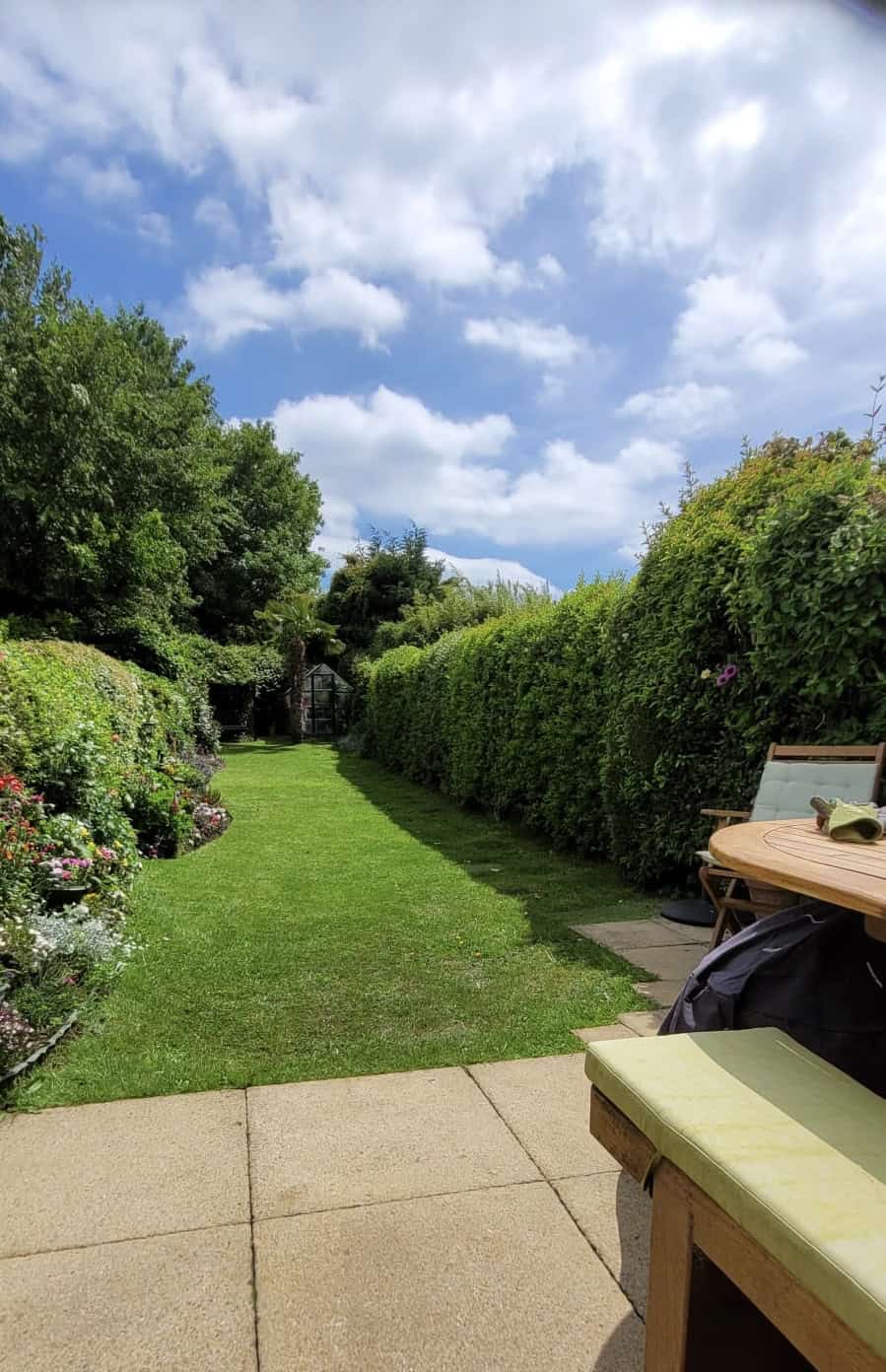 Wide view of Alex’s English cottage garden featuring neatly trimmed evergreen hedges, colourful flower borders, a manicured lawn, and a greenhouse in the distance.