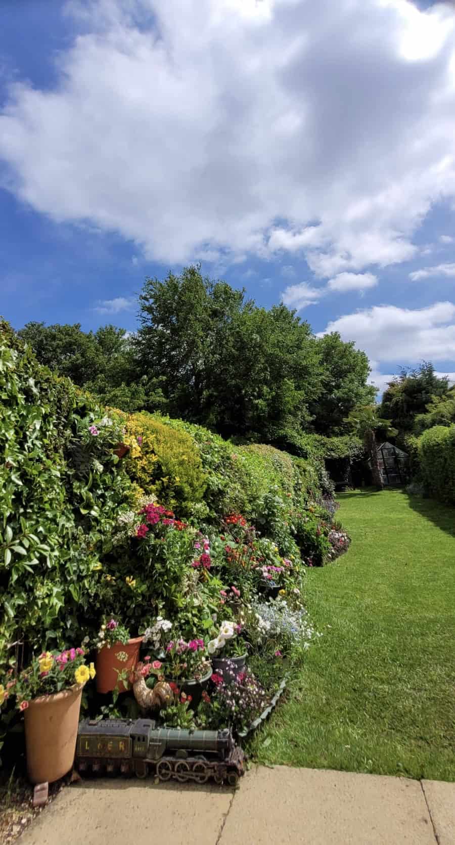 Beautiful English cottage garden with colourful flower borders, neatly trimmed evergreen hedges, a lush green lawn, and a charming greenhouse in the distance.