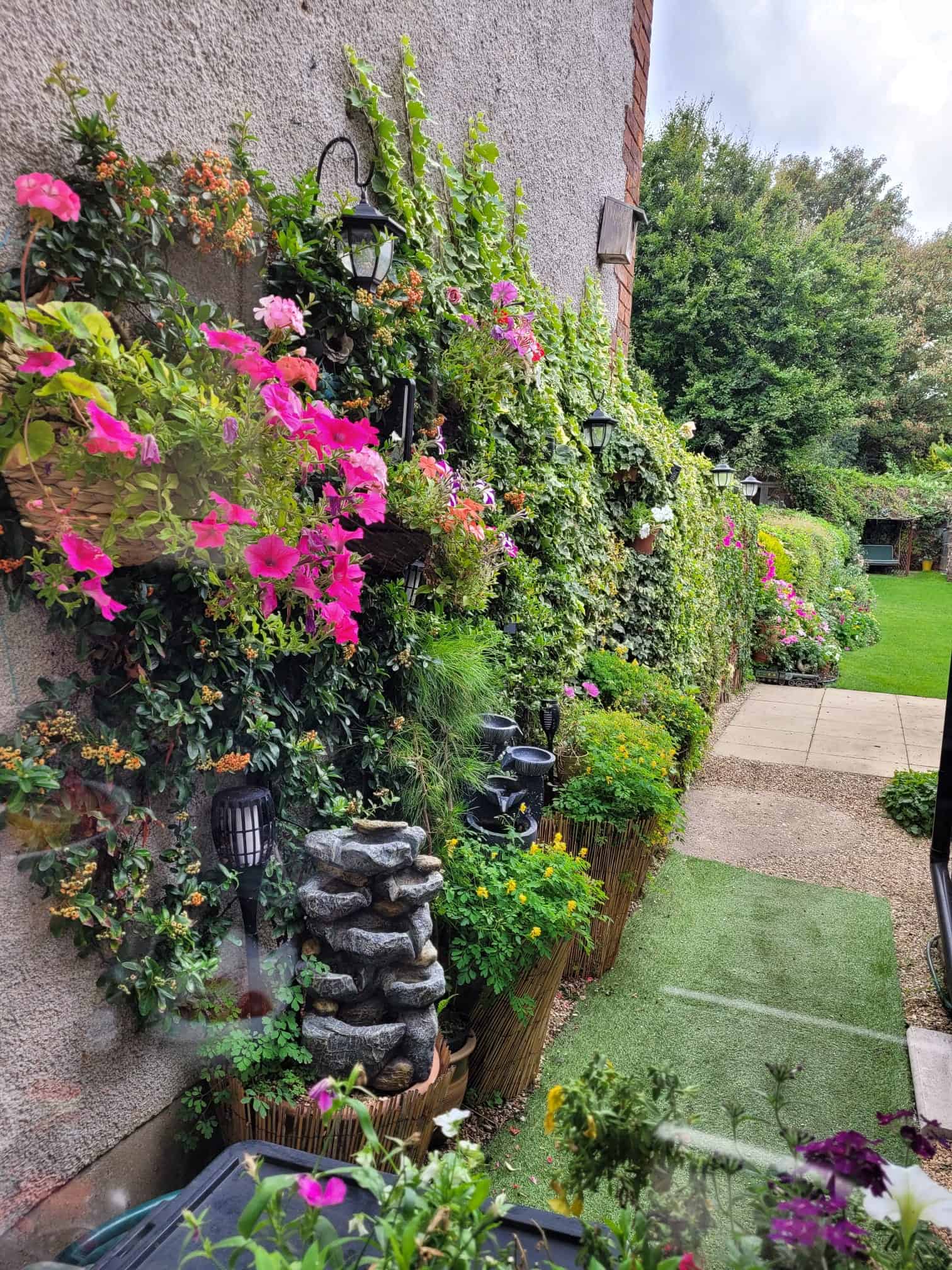 Charming English cottage garden pathway lined with colourful hanging baskets, climbing ivy, flowering plants, decorative lanterns, and a tranquil water feature.