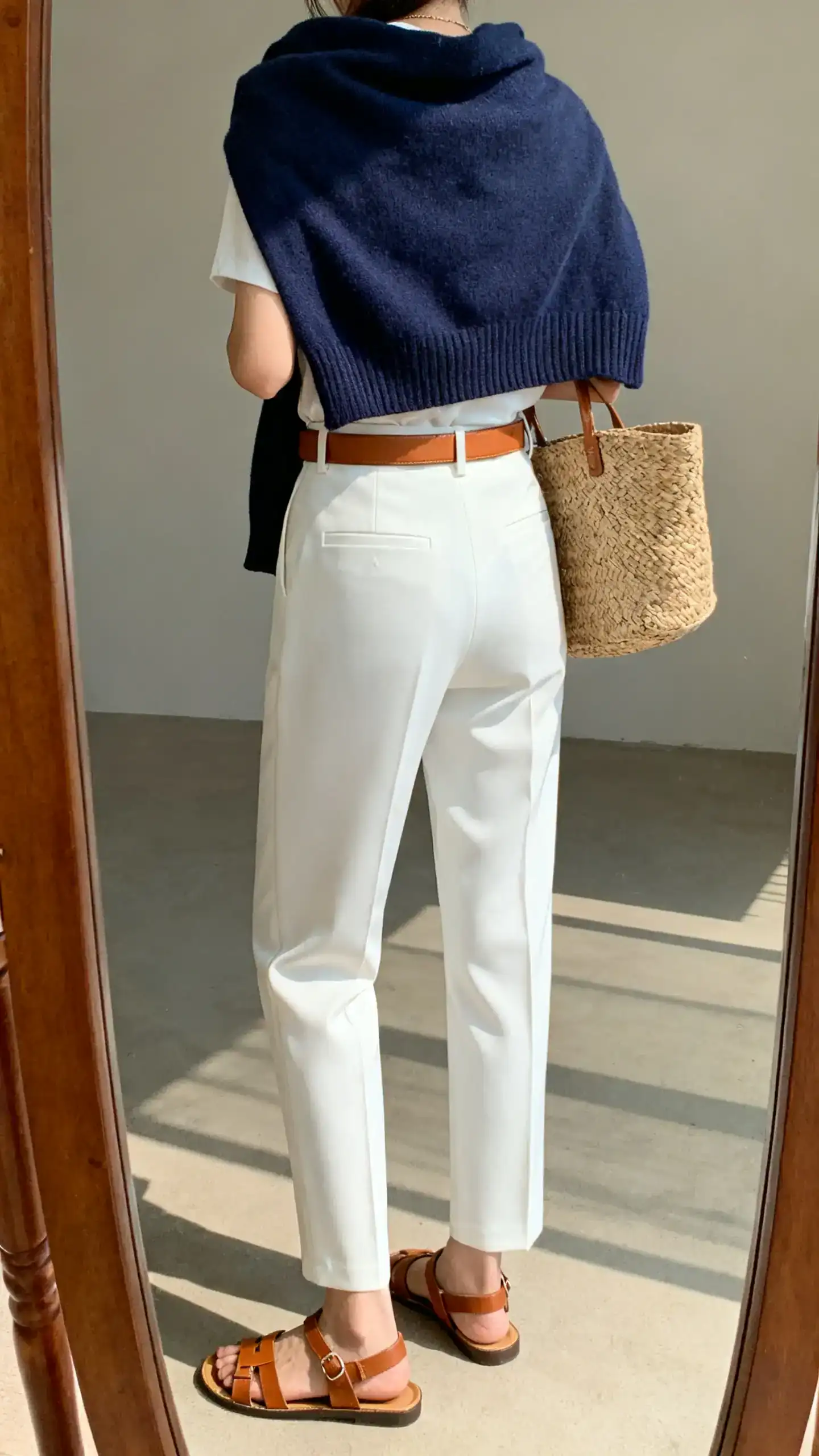 iPhone back view of a woman wearing white tailored trousers with a navy knit sweater draped over the shoulders (or worn), brown leather sandals, a tan belt, and a woven tote, face not visible, full-length mirror, bright natural daylight, iPhone photo quality.