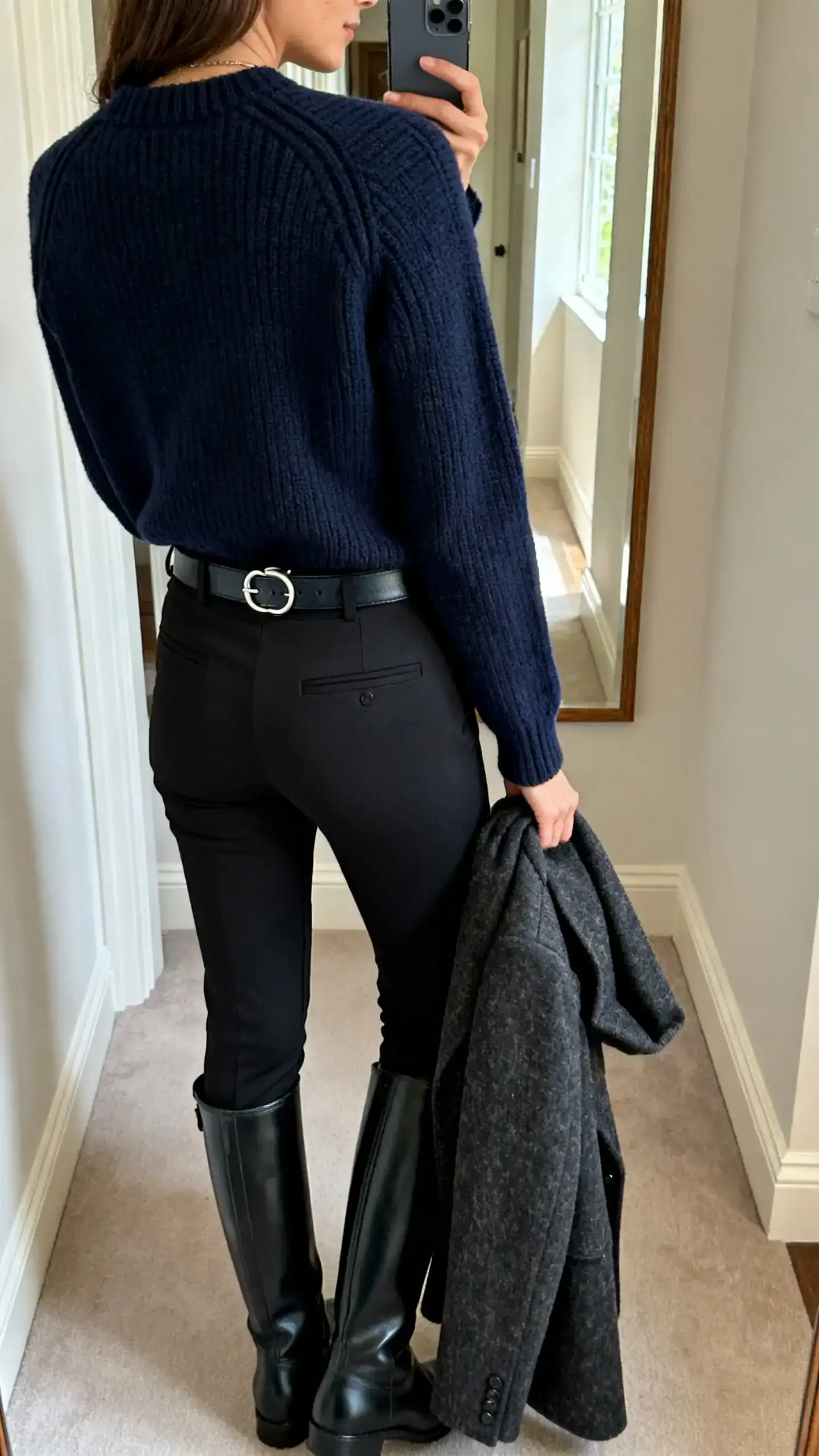 iPhone back-view selfie of a woman in a navy structured knit sweater with slim black tailored leggings, knee-high black riding boots, belt with subtle equestrian buckle, wool coat over arm, face not visible, hallway mirror, natural daylight, iPhone photo quality.