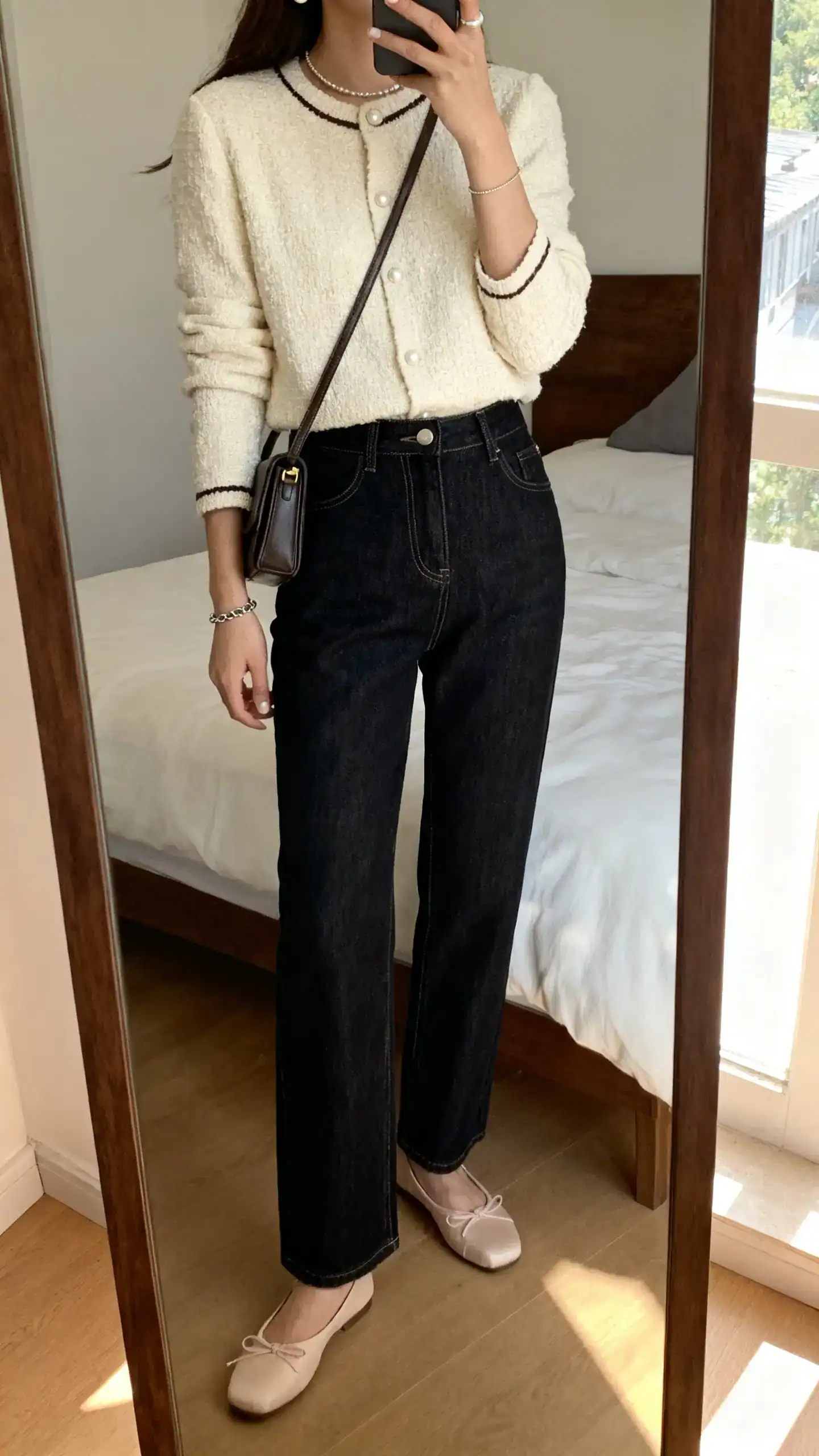 iPhone mirror selfie of a woman in a cream tweed cardigan with minimal trim, high-waisted straight dark-wash jeans, ballet flats, small structured crossbody, pearl studs and slim chain bracelet, face not visible, bedroom mirror, natural afternoon light, iPhone photo quality.