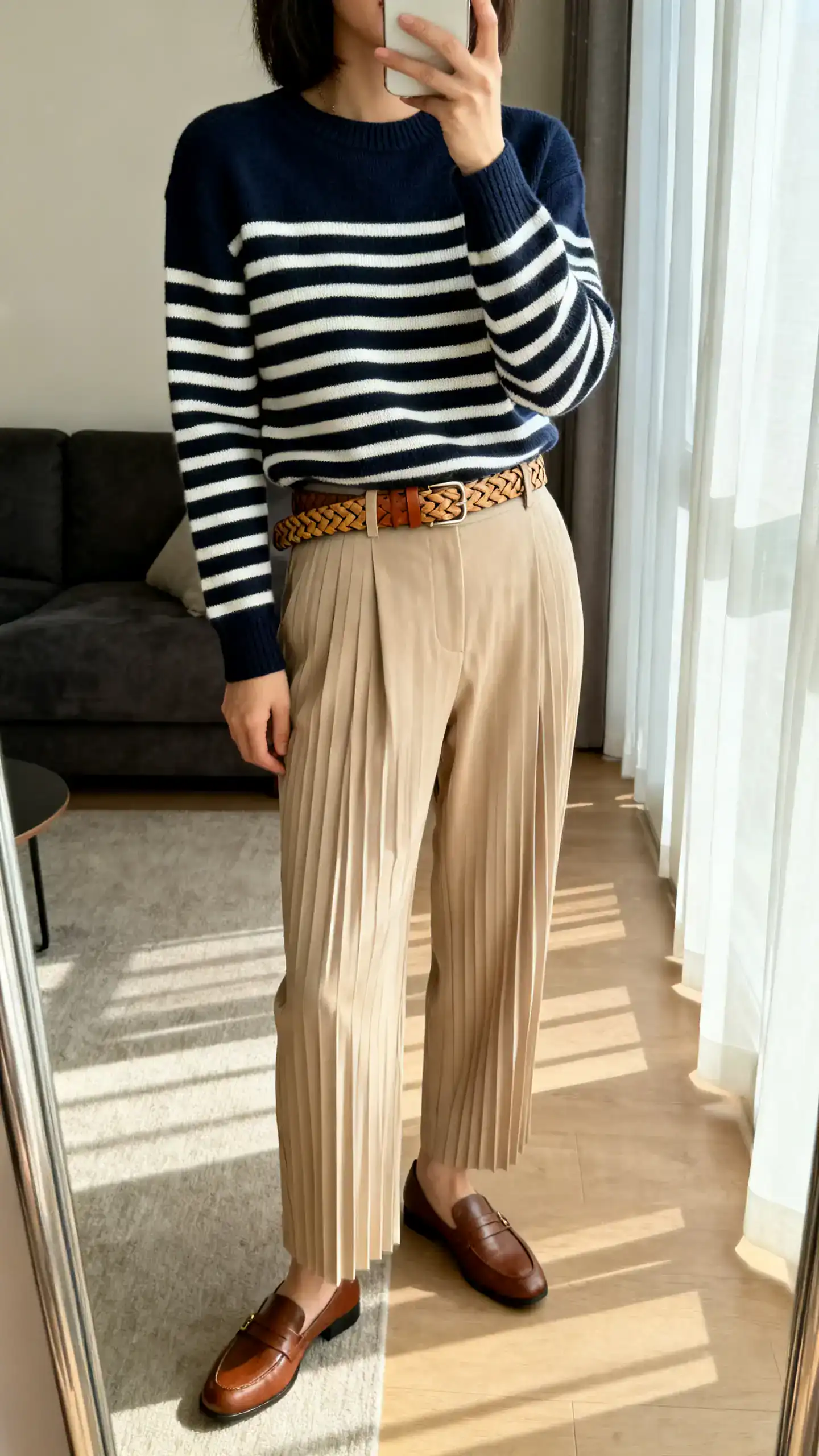 iPhone mirror selfie of a woman wearing a classic Breton stripe knit (navy/cream) with high-waisted pleated beige trousers, brown loafers, and a woven leather belt, face not visible, side angle, living room mirror, morning window light, iPhone photo quality.